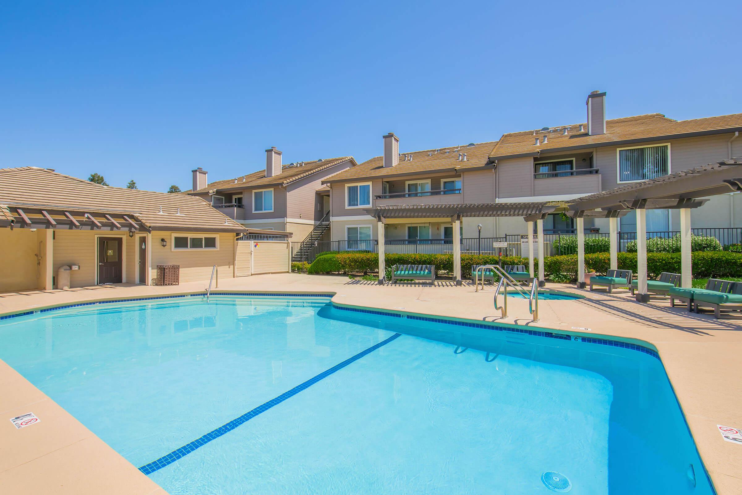 A clear blue swimming pool surrounded by lounge chairs and shaded areas, with two multi-story residential buildings in the background. The sky is bright and sunny, creating a relaxing outdoor atmosphere.