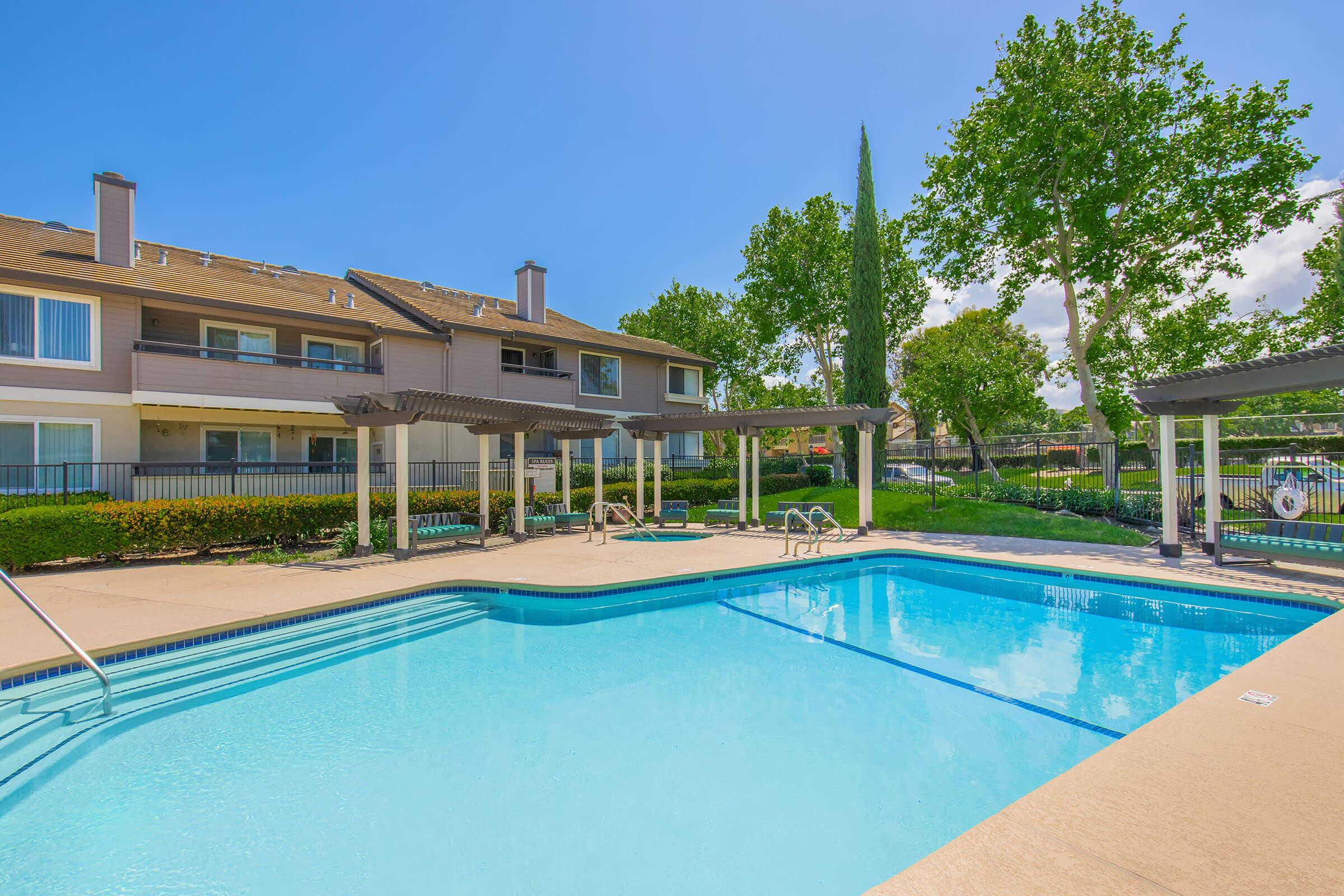 A clear, inviting swimming pool surrounded by lounge chairs and shaded areas. Lush trees and greenery are visible in the background, alongside two multi-story buildings with balconies. The sky is bright blue with a few fluffy clouds, suggesting a warm, sunny day.