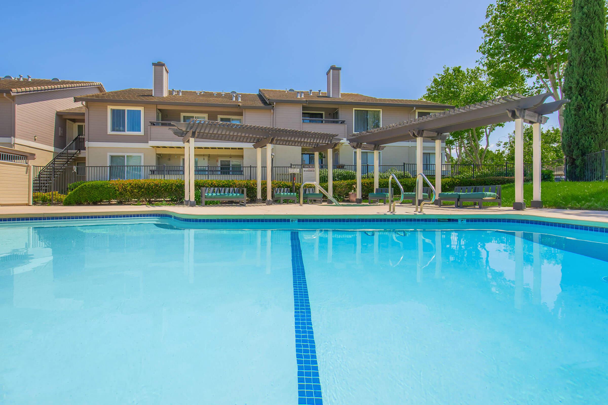 A clear blue swimming pool in the foreground, surrounded by lounge chairs. In the background, two-story apartment buildings with balconies are visible, along with well-maintained landscaping and trees under a bright blue sky. The scene conveys a relaxing outdoor atmosphere.