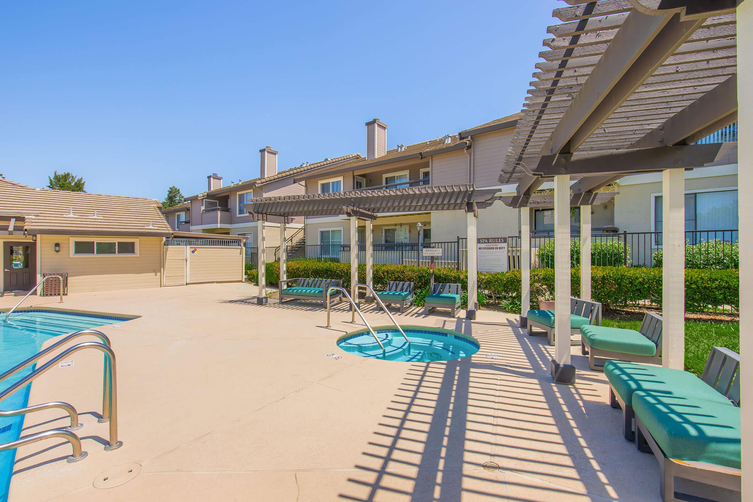 A bright outdoor pool area featuring a main swimming pool, a hot tub, and lounge seating under shaded pergolas. Surrounding greenery adds a fresh touch, with residential buildings in the background and clear blue skies above. The space is designed for relaxation and enjoyment.