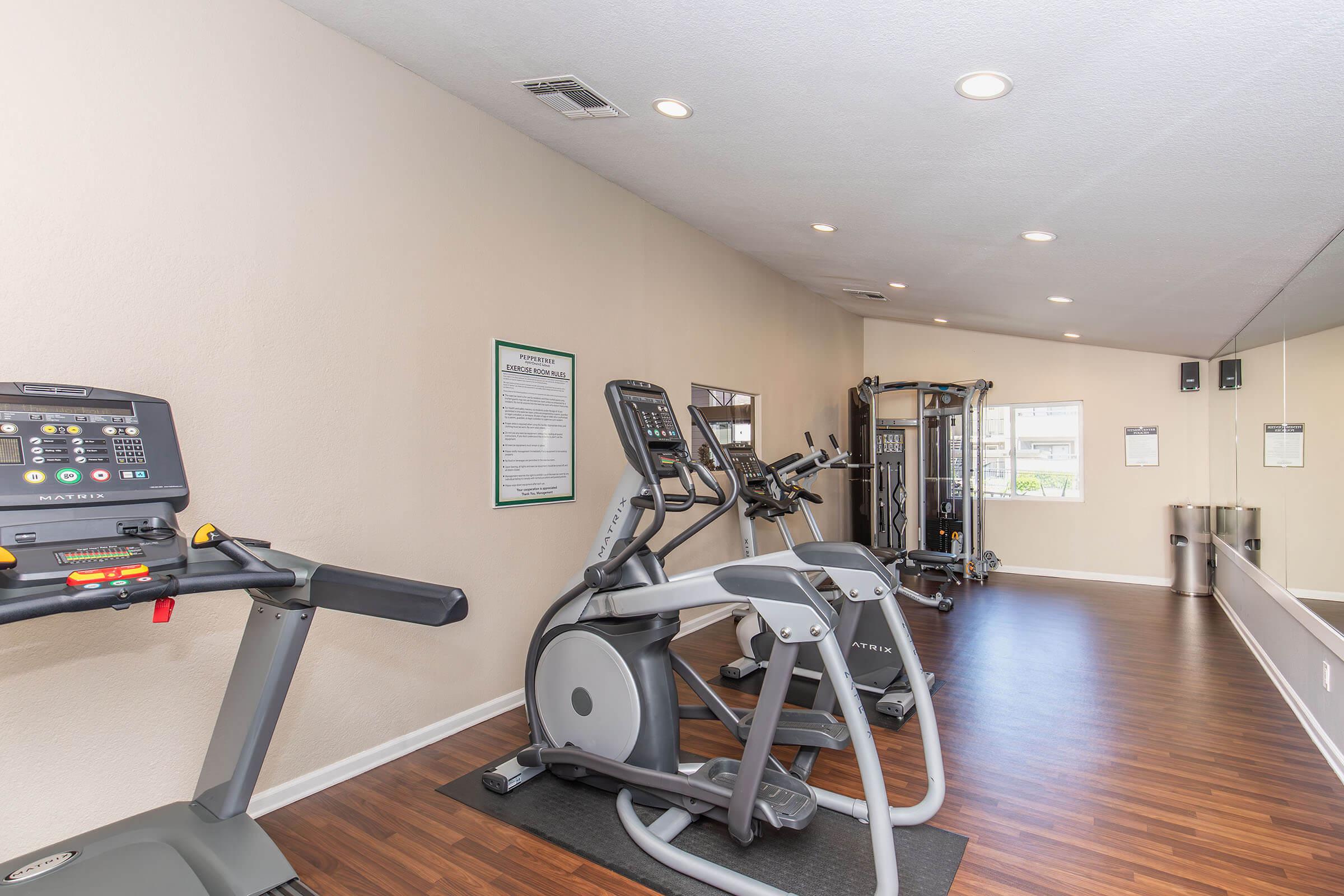 A well-lit fitness room featuring various exercise equipment, including a treadmill, an elliptical machine, and a multi-station gym. The walls are painted a light color, and there is a large mirror reflecting the setup. Wooden flooring adds warmth to the space.
