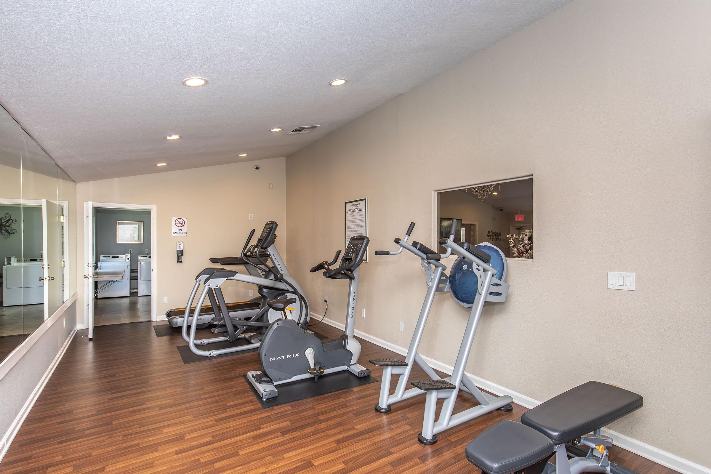 A well-lit gym area featuring exercise equipment, including an elliptical machine, a stationary bike, and a weight bench. The walls are painted a light color, with a large mirror and informational posters visible. The flooring is dark wood, creating a modern and inviting atmosphere for workouts.