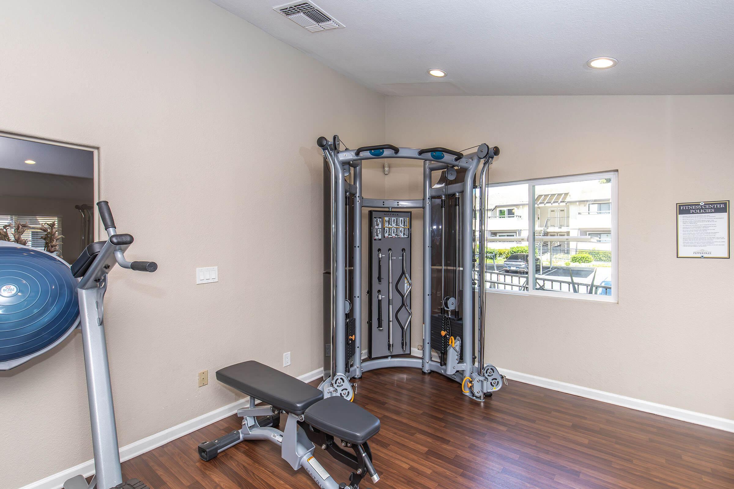 A modern gym interior featuring a multi-station workout machine, a weight bench, and a stationary bike. Natural light enters through a window, illuminating the wooden floor and beige walls, creating a spacious and inviting workout environment.