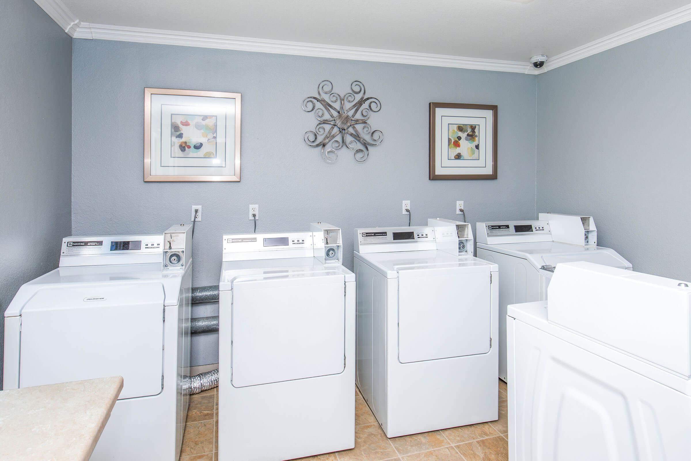A clean and modern laundry room featuring four white washing machines arranged in a row. The walls are painted a soft gray, with two framed artworks hanging above the machines. There is a decorative wall piece in the center, adding a stylish touch to the space. The floor is tiled in a light, neutral color.