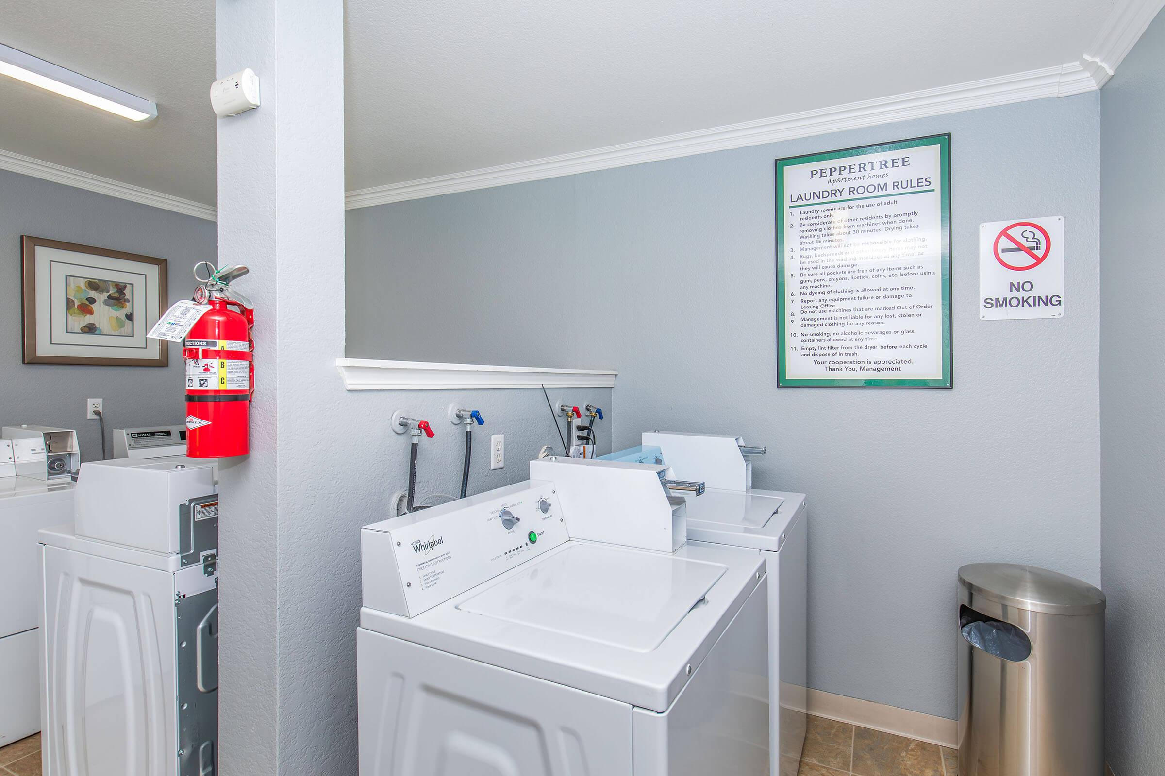 A laundry room featuring several white washing machines and dryers. The walls are painted gray, and there is a fire extinguisher mounted on the wall. A sign detailing laundry room rules is prominently displayed. A trash can is also visible in the corner of the room. No smoking sign is posted nearby.