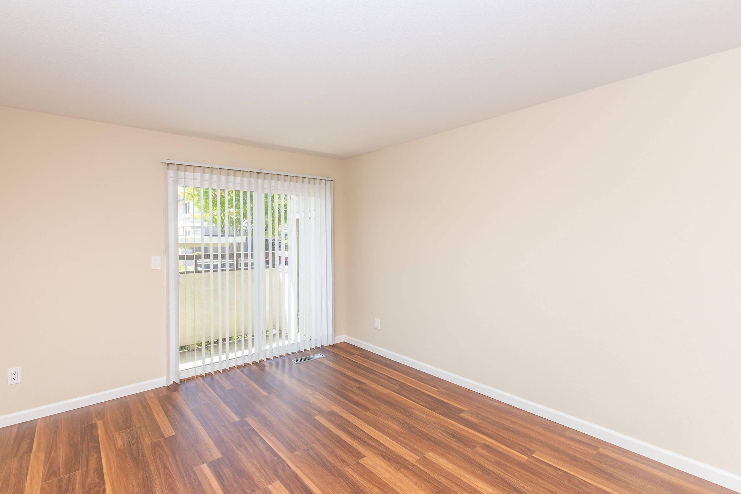 A bright, empty room featuring light-colored walls and wooden flooring. A sliding glass door with vertical blinds allows natural light to enter, leading to an outdoor space. The room is devoid of furniture, creating a minimalist ambiance.