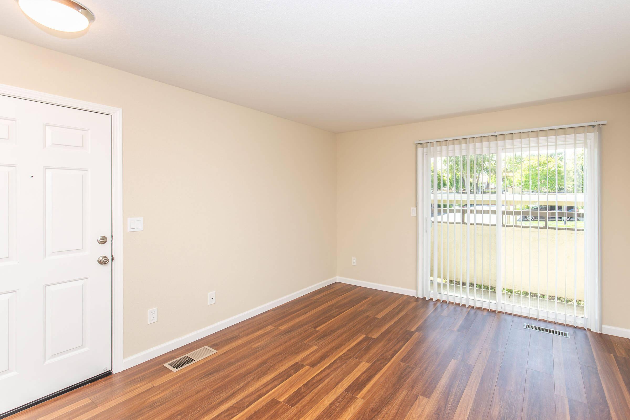 A well-lit interior room with light beige walls and hardwood flooring. The space features a white front door on the left and a large window with vertical blinds on the right, allowing natural light to enter. The room is empty, providing a blank canvas for potential furniture arrangements and decor.