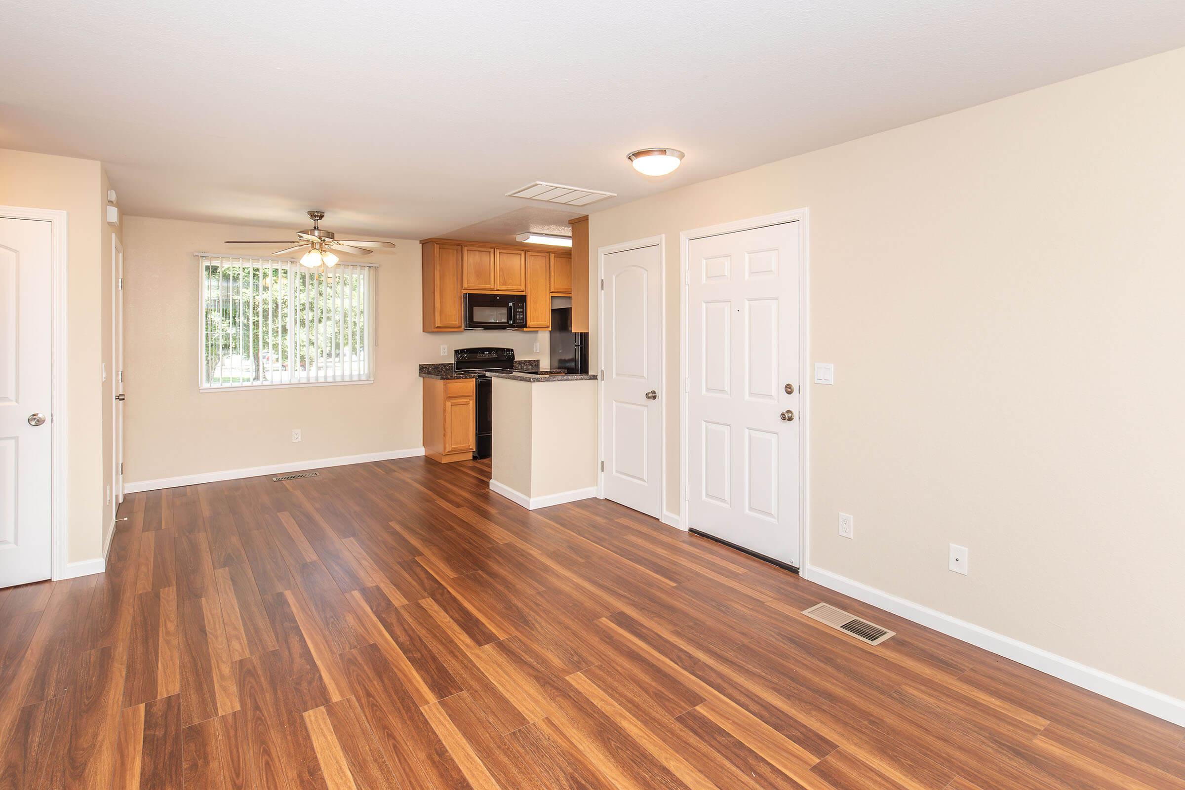 Spacious living area featuring hardwood floors, a ceiling fan, and natural light from a window. The kitchen area includes wooden cabinets and modern appliances. Two doors lead to separate rooms, and there's a front door visible, enhancing access and flow in this inviting open layout.