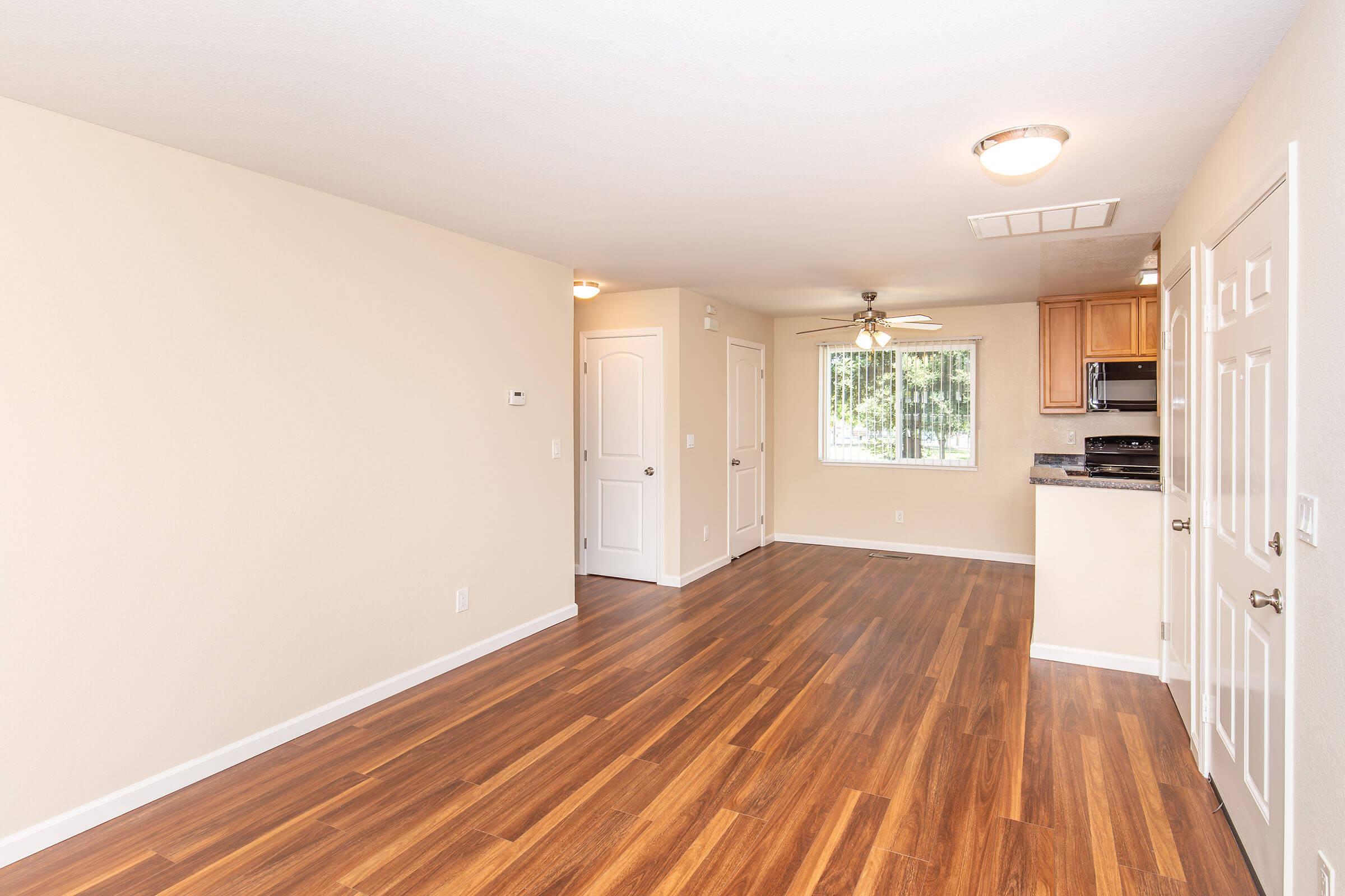 An interior view of a modern, unfurnished living space featuring light beige walls and dark wooden flooring. A window allows natural light into the room, which includes a ceiling fan. The kitchen area is visible in the background, with cabinets and appliances shown.