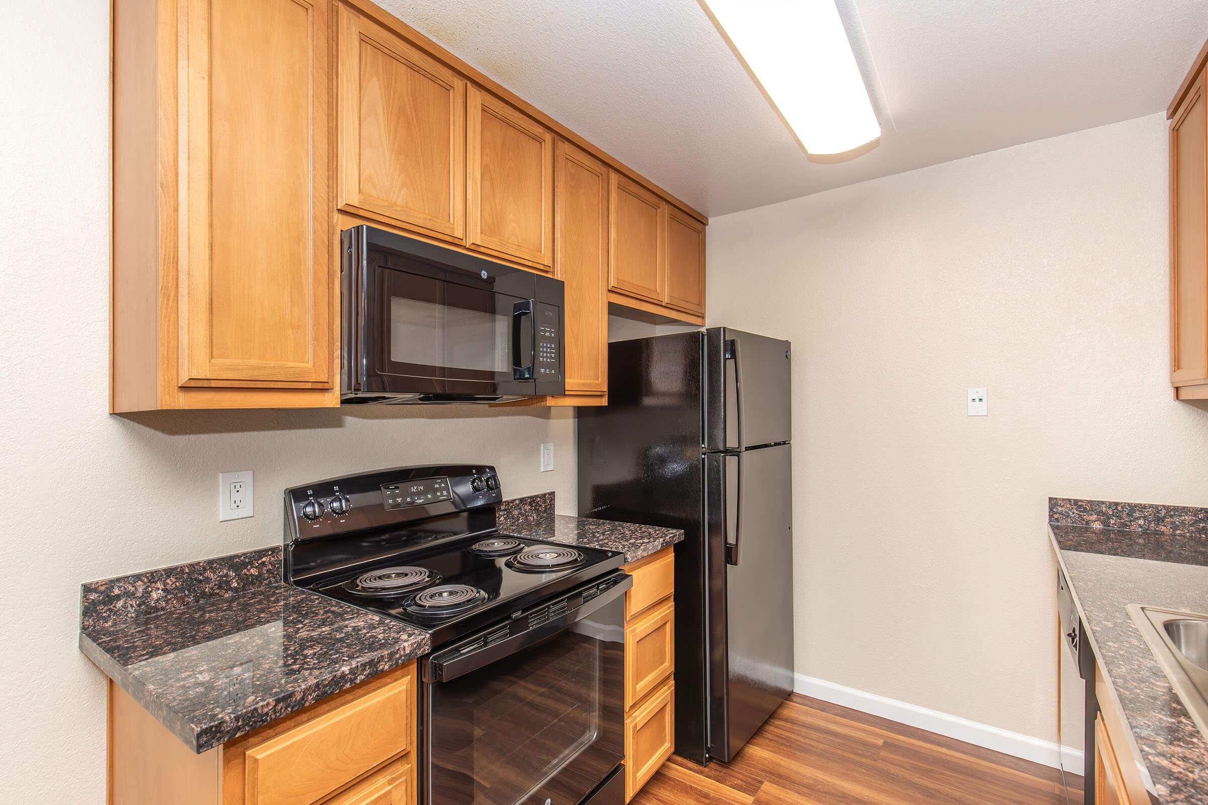 A modern kitchen featuring wooden cabinets, a black refrigerator, a black microwave above a stove, and granite countertops. The space is brightly lit with a ceiling light, and the walls are painted a light color, creating a clean and inviting atmosphere.