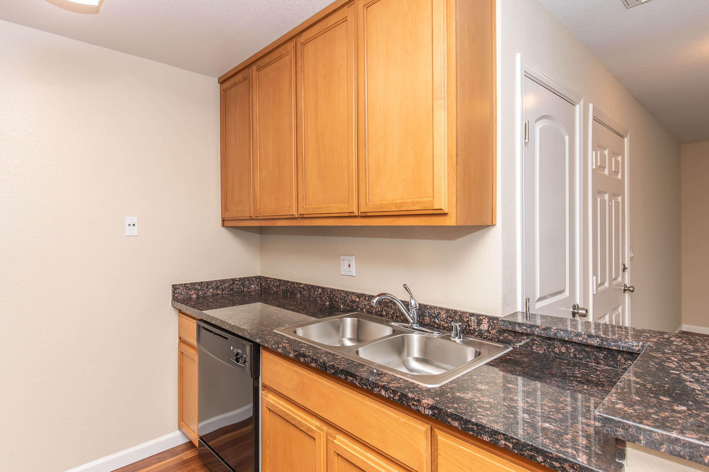 A modern kitchen featuring wooden cabinets, a granite countertop with a double sink, a stainless steel dishwasher, and light-colored walls. The kitchen has an open layout leading to a doorway, providing a spacious and inviting atmosphere.
