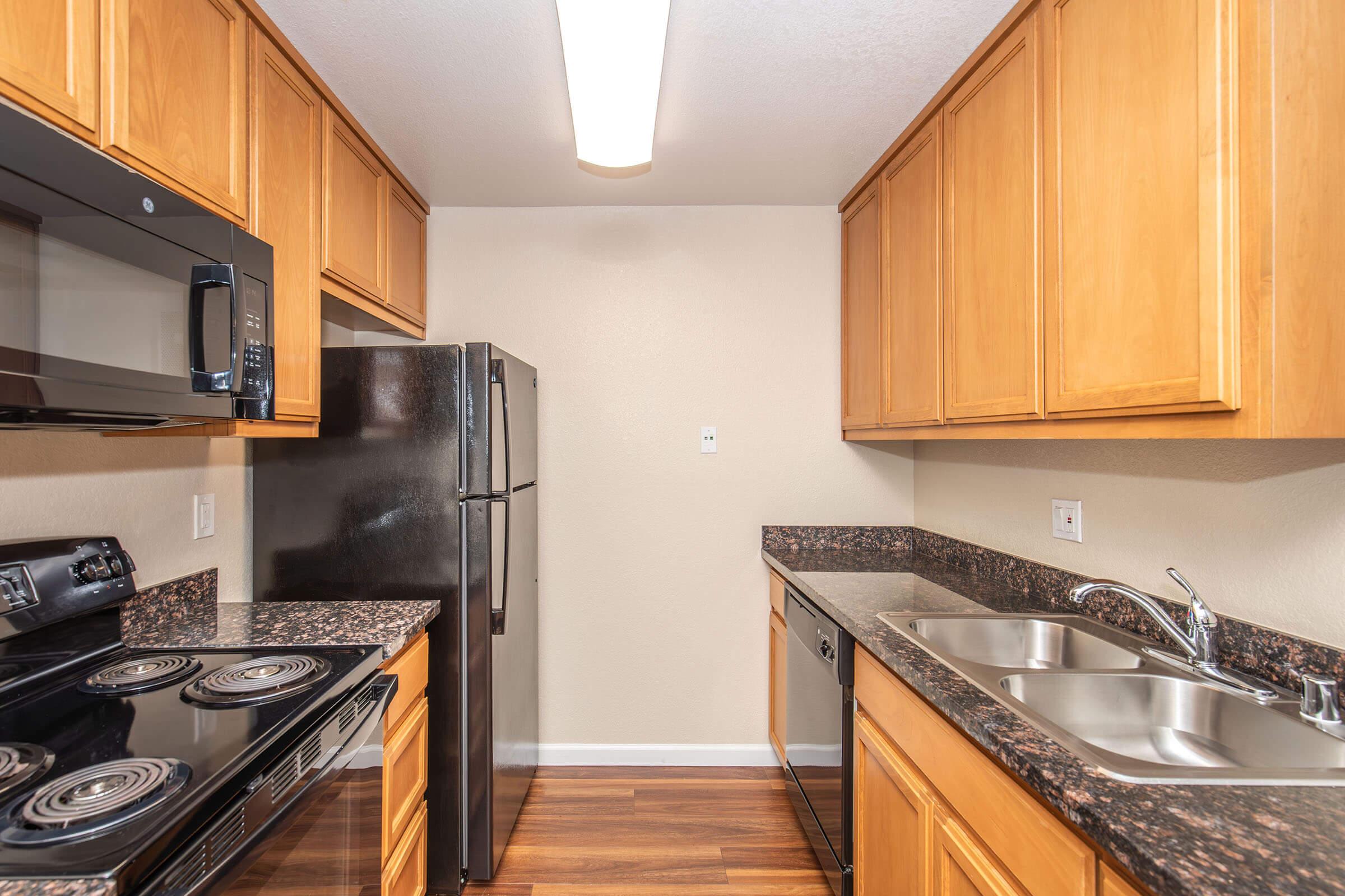 A modern kitchen featuring light wood cabinets, a black microwave, a black refrigerator, a dark granite countertop, and stainless steel sinks. The layout includes an electric stove and a dishwasher, with warm lighting and wooden flooring to create a welcoming ambiance.