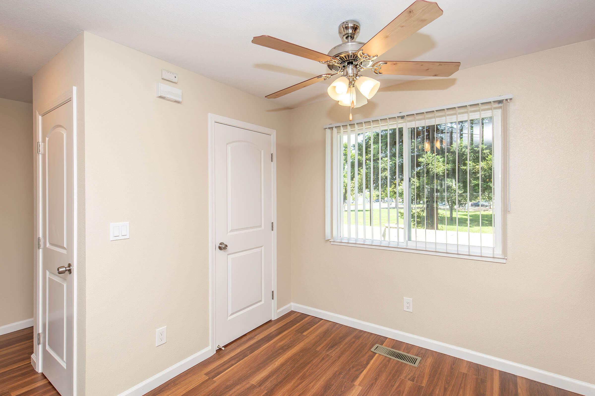 A well-lit room featuring a ceiling fan with lights, beige walls, and wooden flooring. There's a window with vertical blinds providing natural light and a glimpse of greenery outside. A white door leads to another area, enhancing the room's inviting and cozy atmosphere.