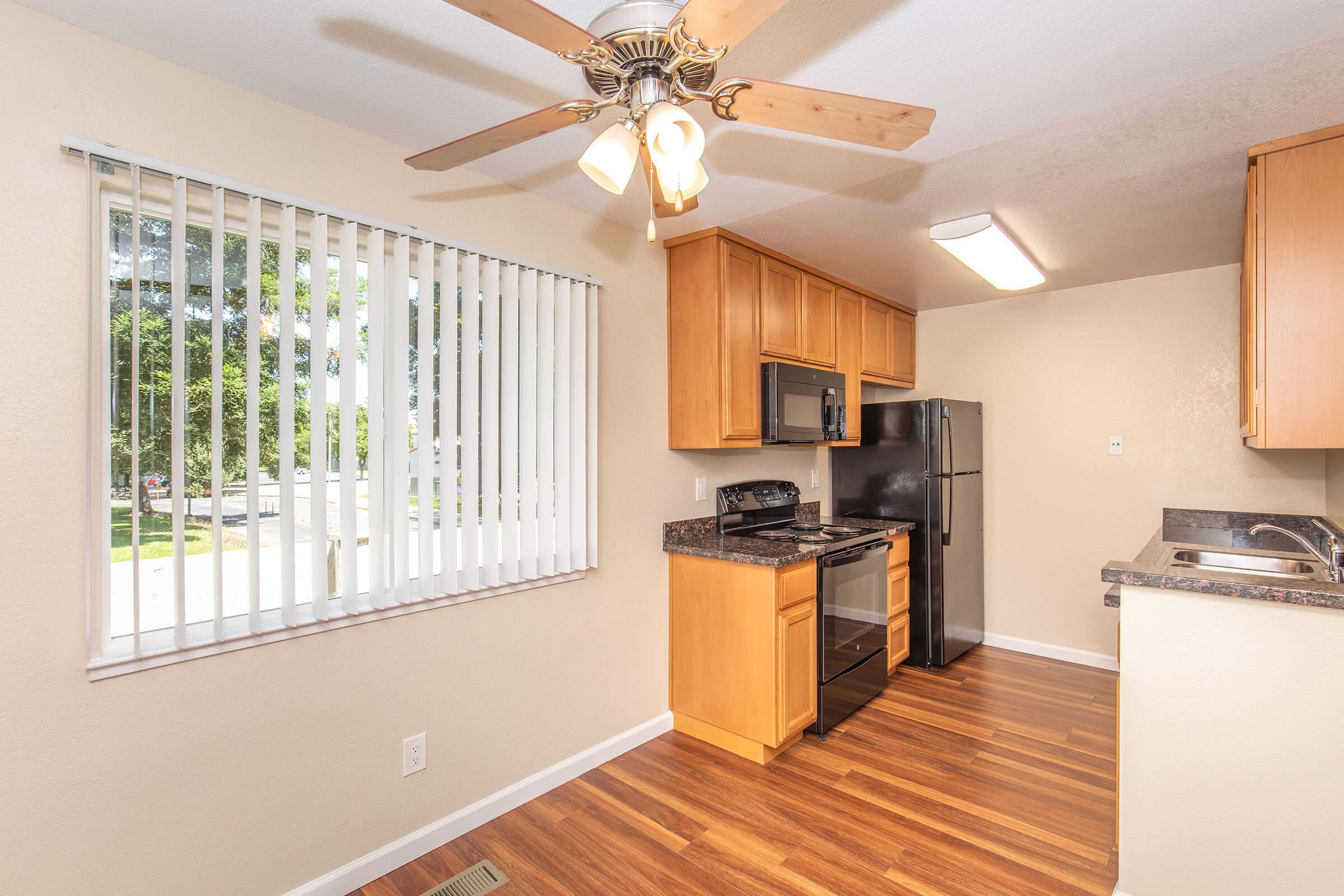 A bright kitchen featuring wooden cabinets, a black refrigerator, and a stove with an oven. There is a ceiling fan with light, and a window with vertical blinds allowing natural light to enter. The flooring is a polished wood, creating a warm and inviting atmosphere.