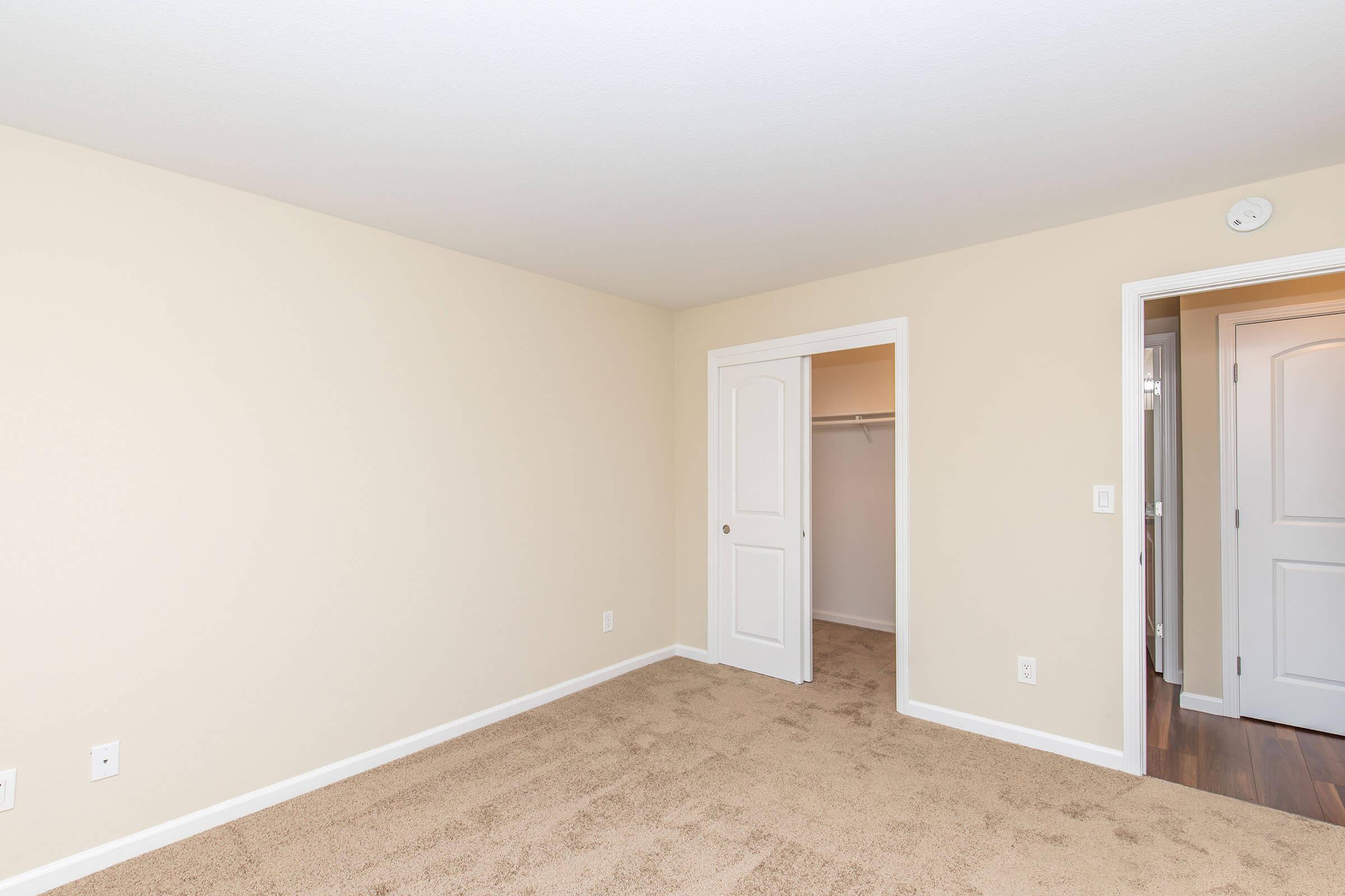 Empty room with tan walls and carpet, featuring a white door and closet. There is an open doorway leading to another room. The space is well-lit and minimalistic, creating a neutral aesthetic suitable for various uses or furnishings.