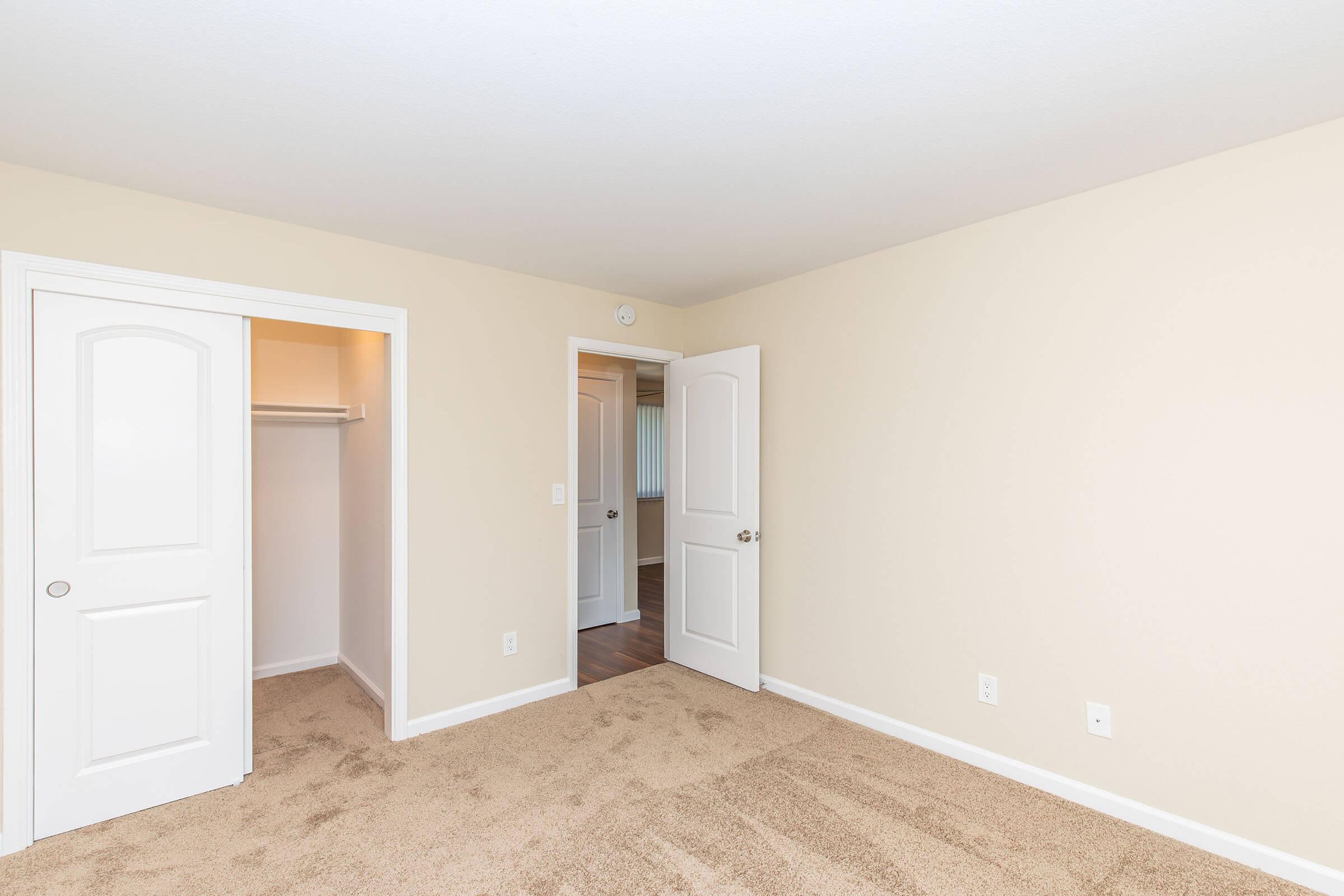 A well-lit, empty bedroom featuring beige walls and carpeted flooring. Two white doors lead to a closet and another room. The space appears clean and inviting, suitable for new occupants to personalize.
