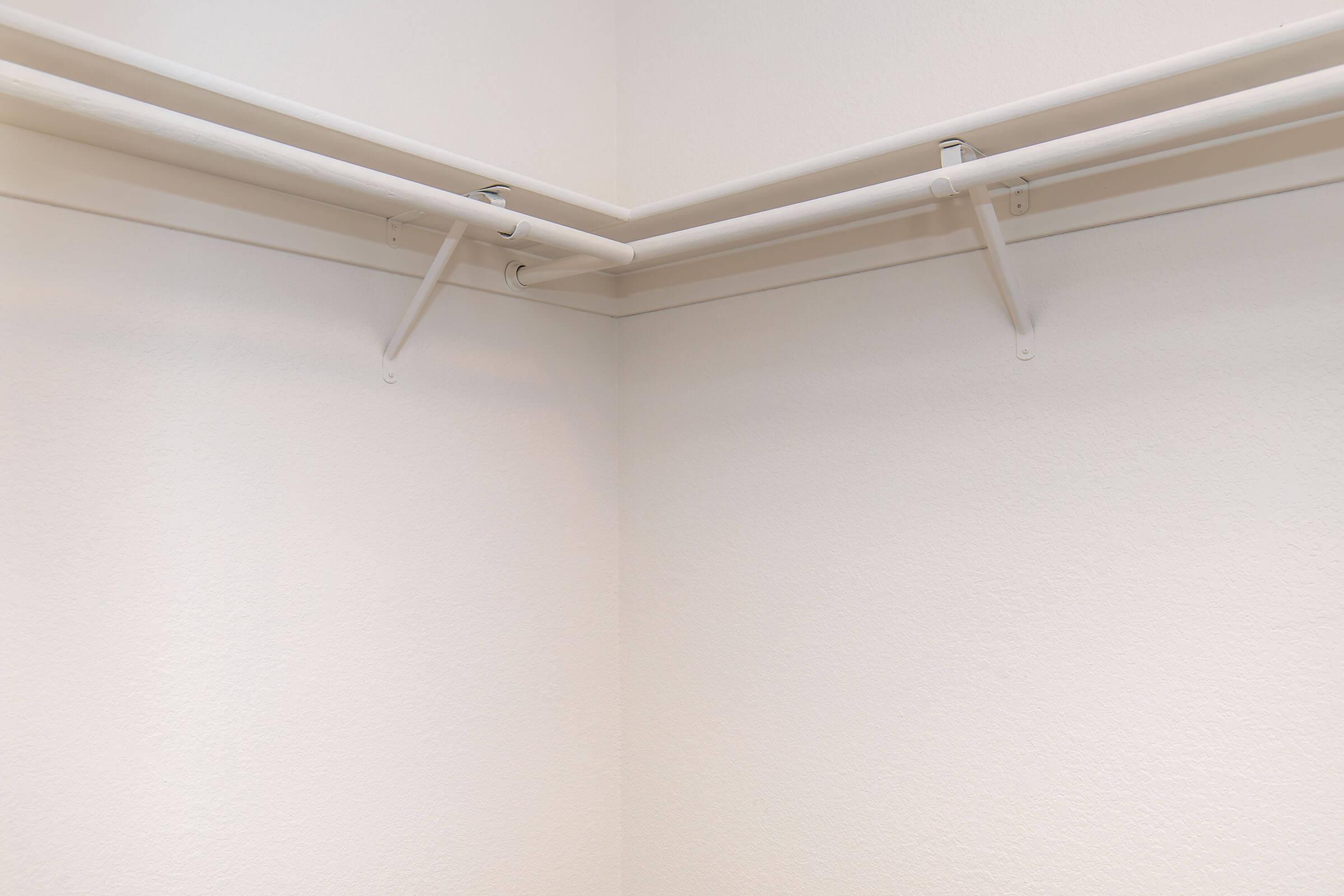 A corner of a closet showing two horizontal white rods mounted on the walls, supported by brackets. The walls are plain and painted in a light color, creating a clean and minimalistic look. No other items are present in the space.