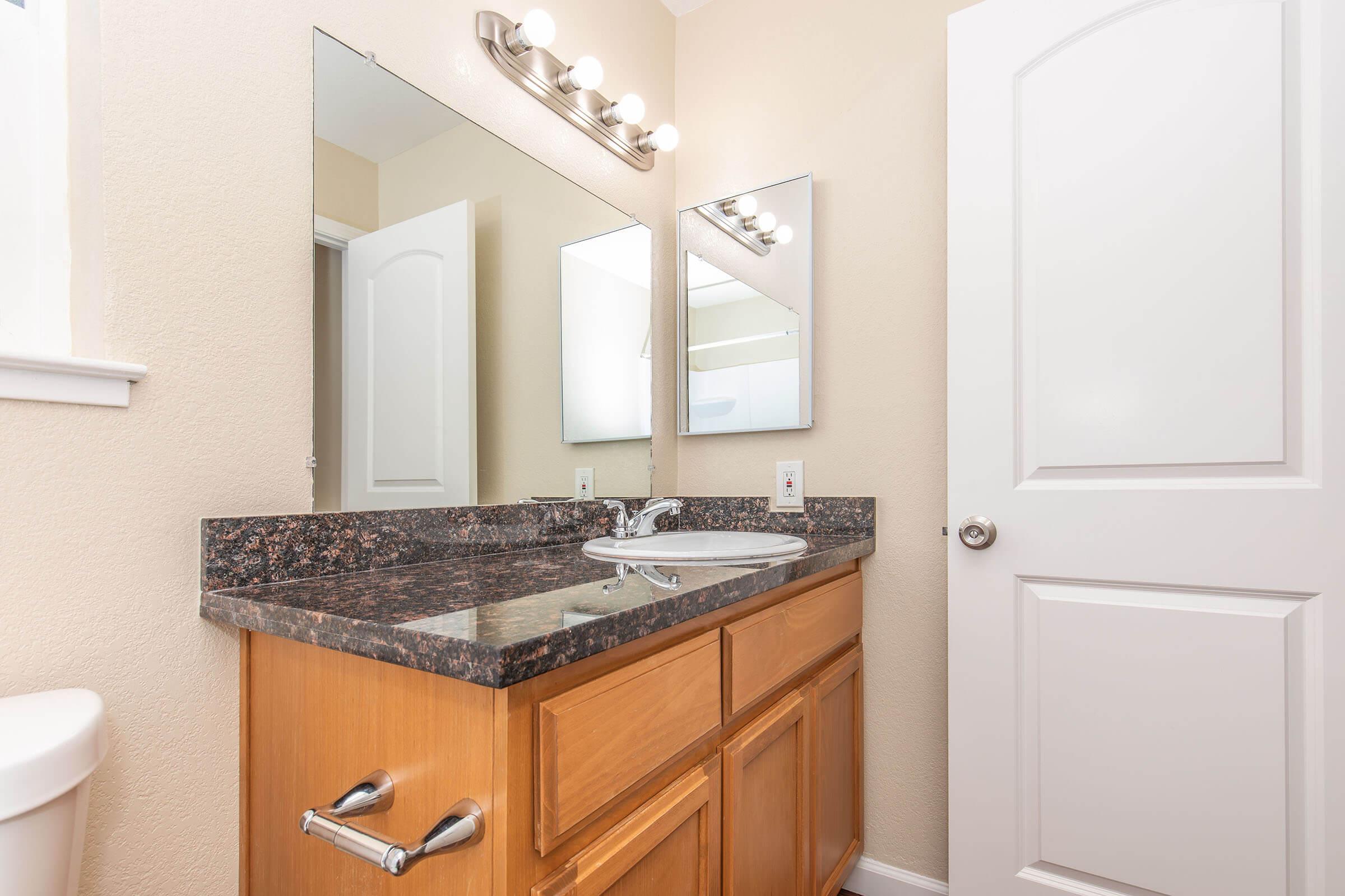 A clean, modern bathroom featuring a dark granite countertop with a sink, a large mirror above, and a wall-mounted light fixture with multiple bulbs. The cabinetry is wooden, and there is a white door leading to another room. A toilet is visible in the background. The walls are painted a light beige color.