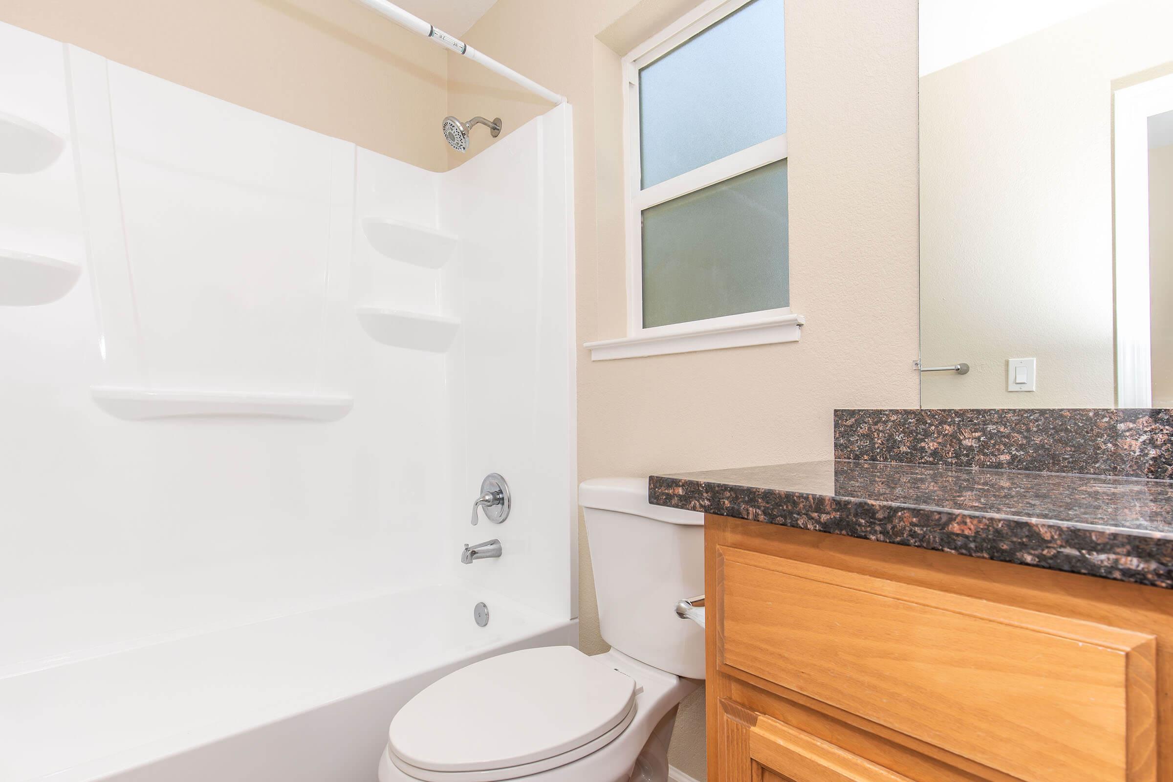 A clean, modern bathroom featuring a white bathtub with a showerhead, a tiled wall, a wooden vanity with a dark countertop, a toilet, and a window allowing natural light. The walls are painted in a light color, creating a bright and airy atmosphere.