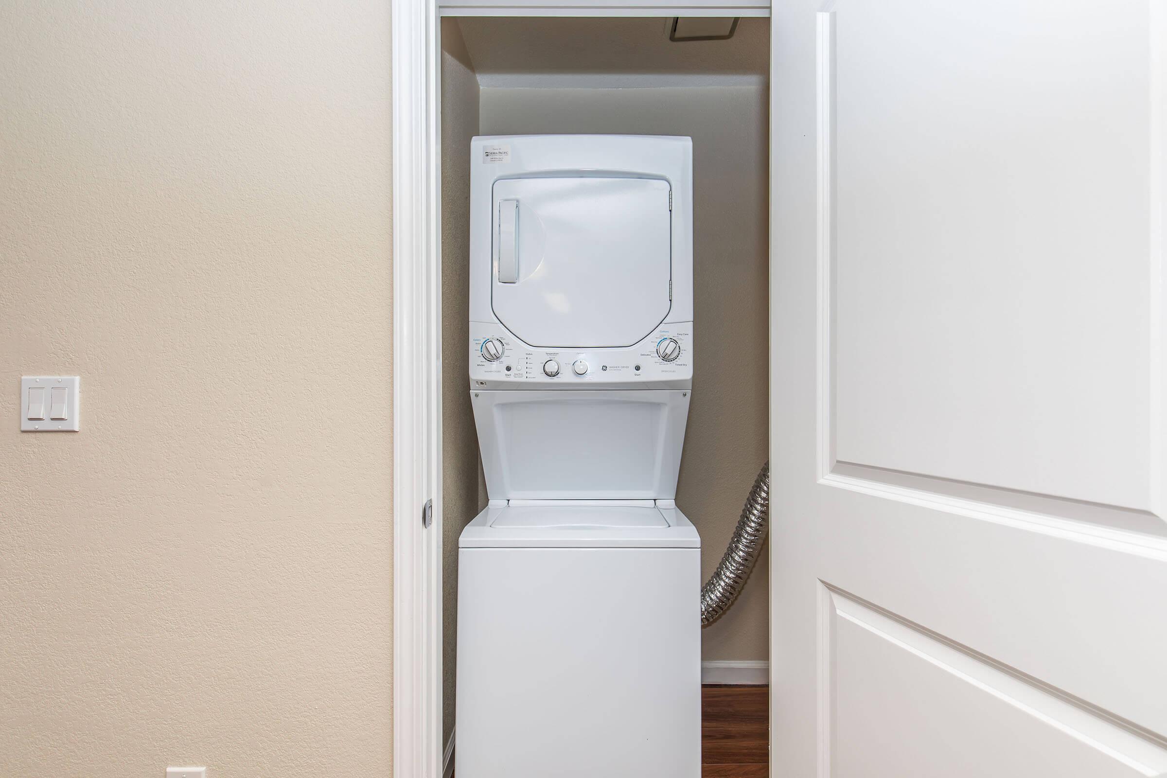 A stacked washer and dryer unit located in a small laundry space, situated between a wall and a closed door. The appliances are white, and the walls are a neutral color, with a vent hose visible behind the dryer. The floor is wooden, and there are no additional items in the laundry area.