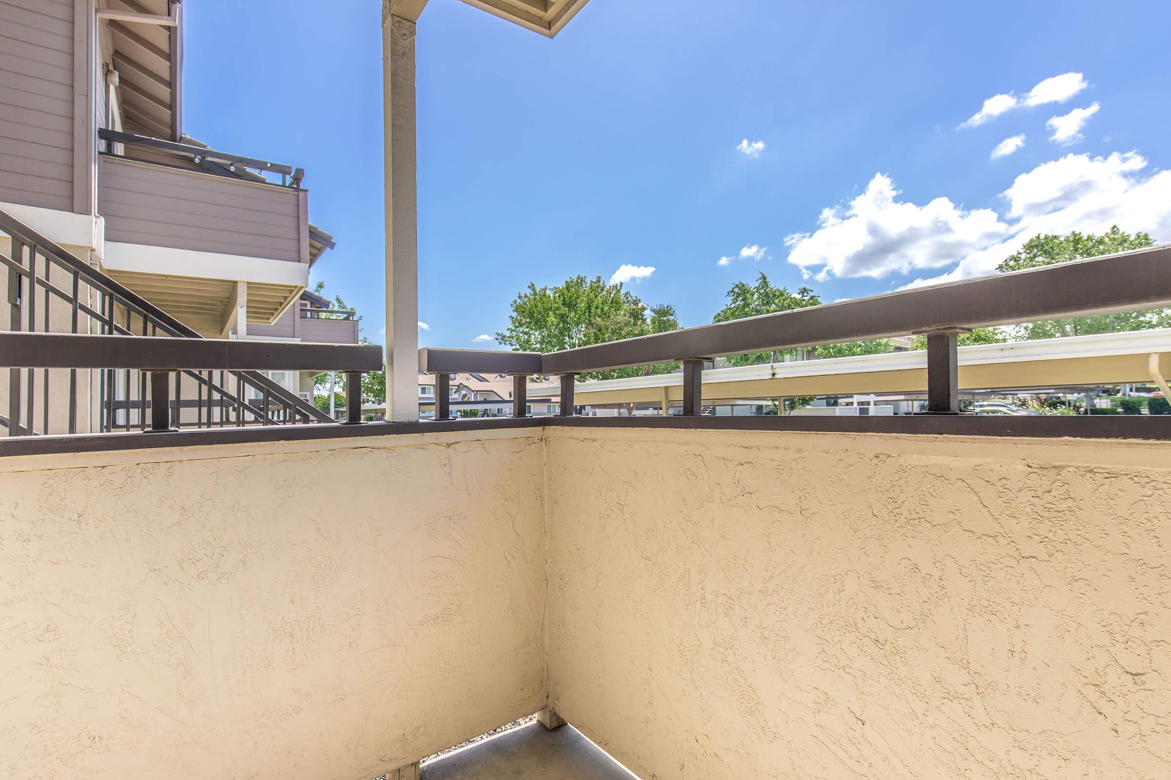 View from a balcony overlooking a sunny outdoor space with blue skies and scattered clouds. The balcony features a beige wall and a wooden railing, with trees visible in the distance. The scene suggests a serene, residential environment.