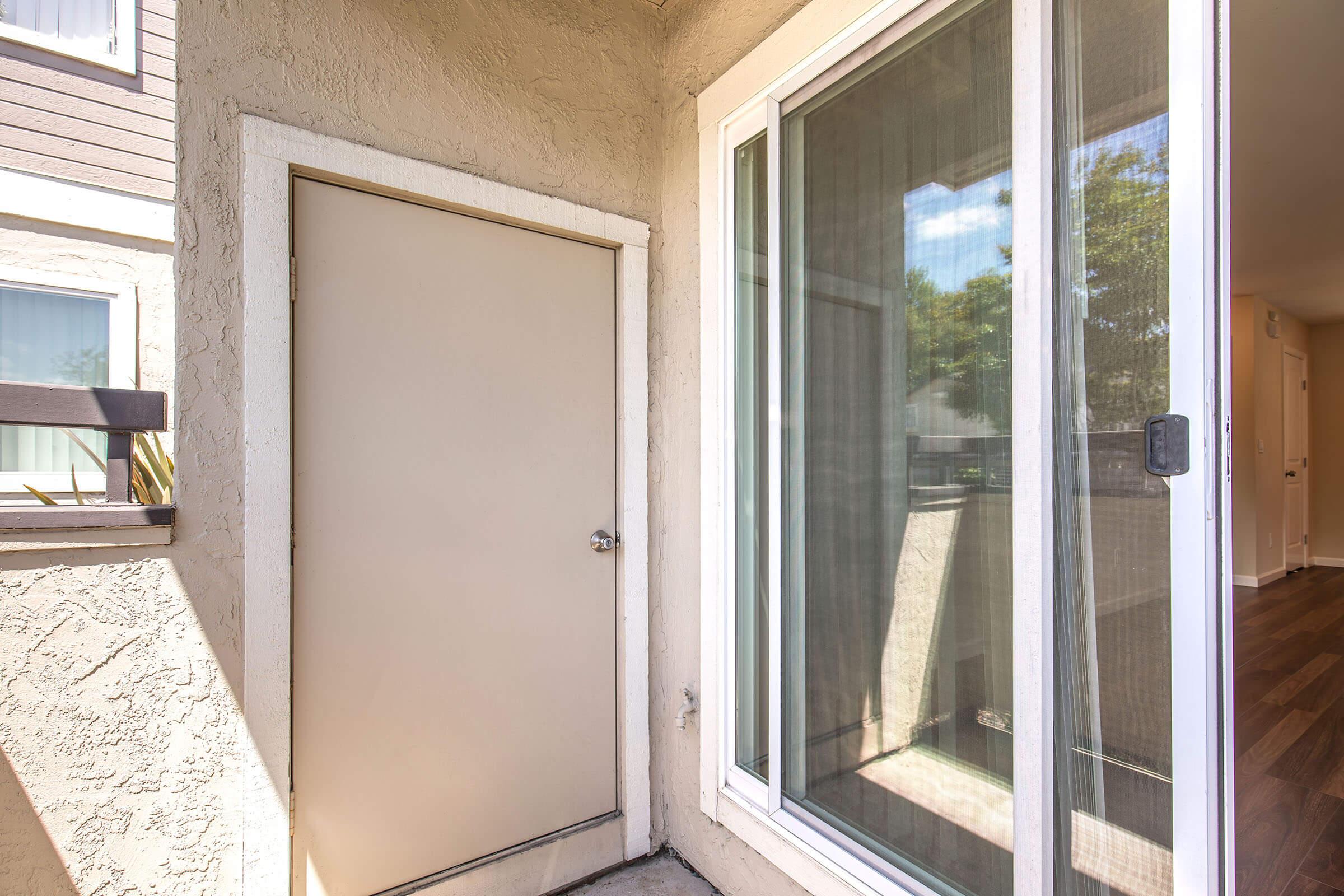A view of a corner entryway featuring a beige door on the left, adjacent to large glass sliding doors. Natural light filters in, illuminating the area. The walls are textured and painted in a soft color, with a glimpse of greenery outside seen through the sliding doors.