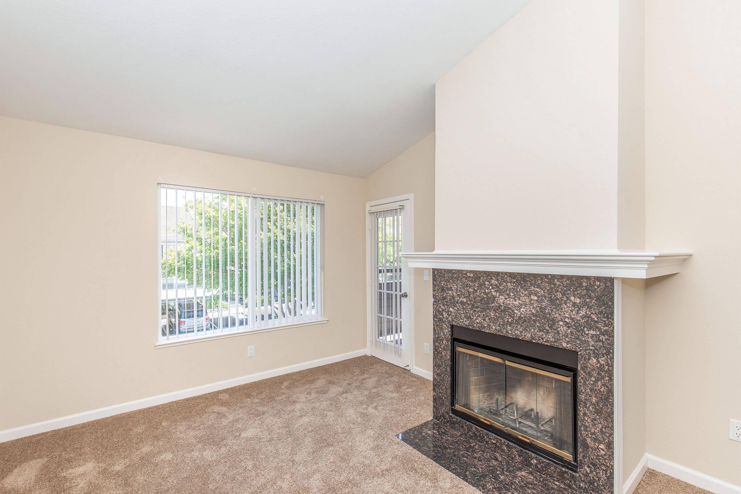 A spacious living room featuring a beige carpet, a decorative fireplace with a marble surround, and large windows with vertical blinds. There is a door leading outside, allowing natural light to illuminate the space. The walls are painted a light color, creating a warm and inviting atmosphere.
