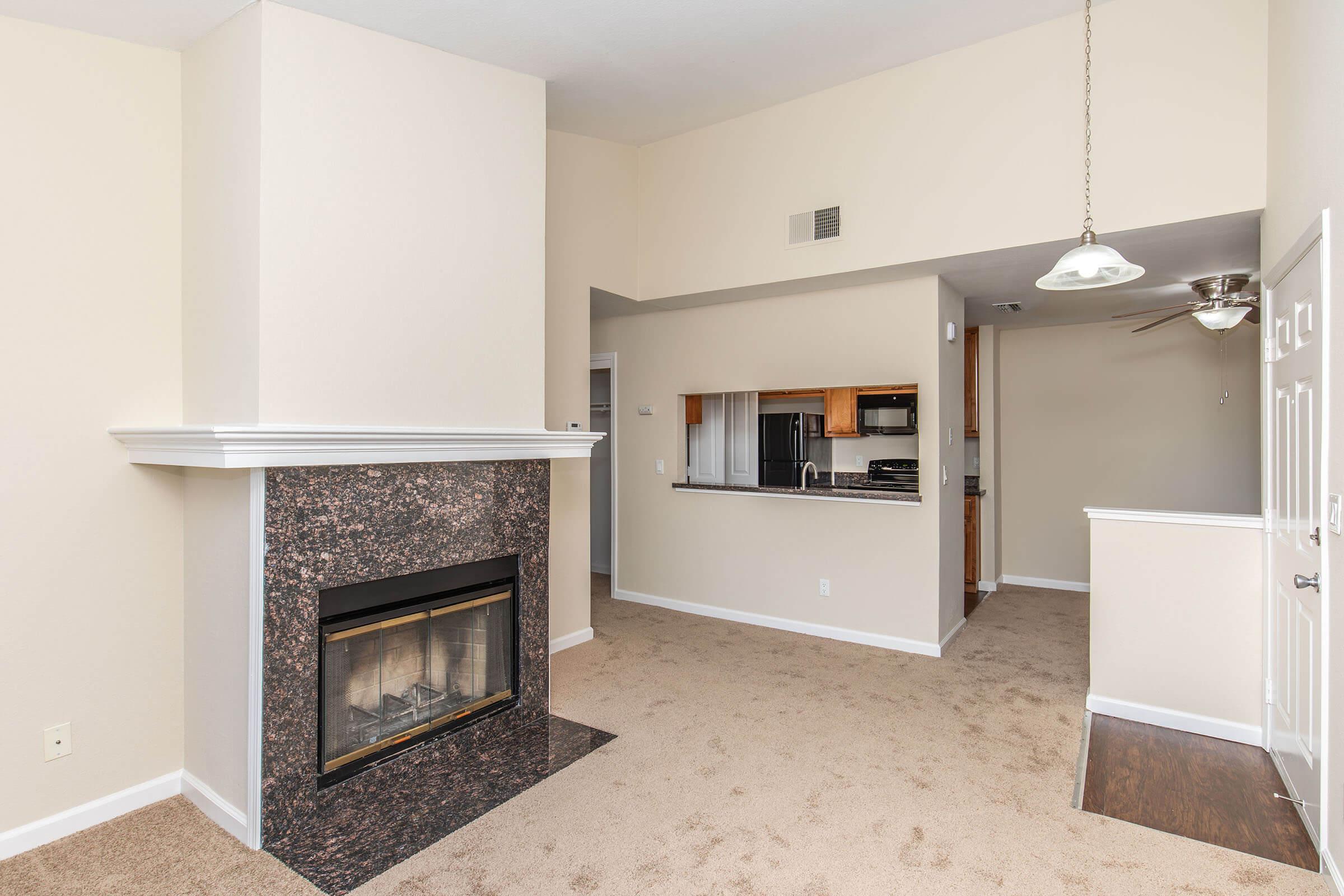 A living room with beige walls and carpet, featuring a cozy fireplace with a dark stone surround. An open kitchen view with wooden cabinets and appliances is visible in the background. There is a ceiling fan above, and a door leading to another room or area can be seen.