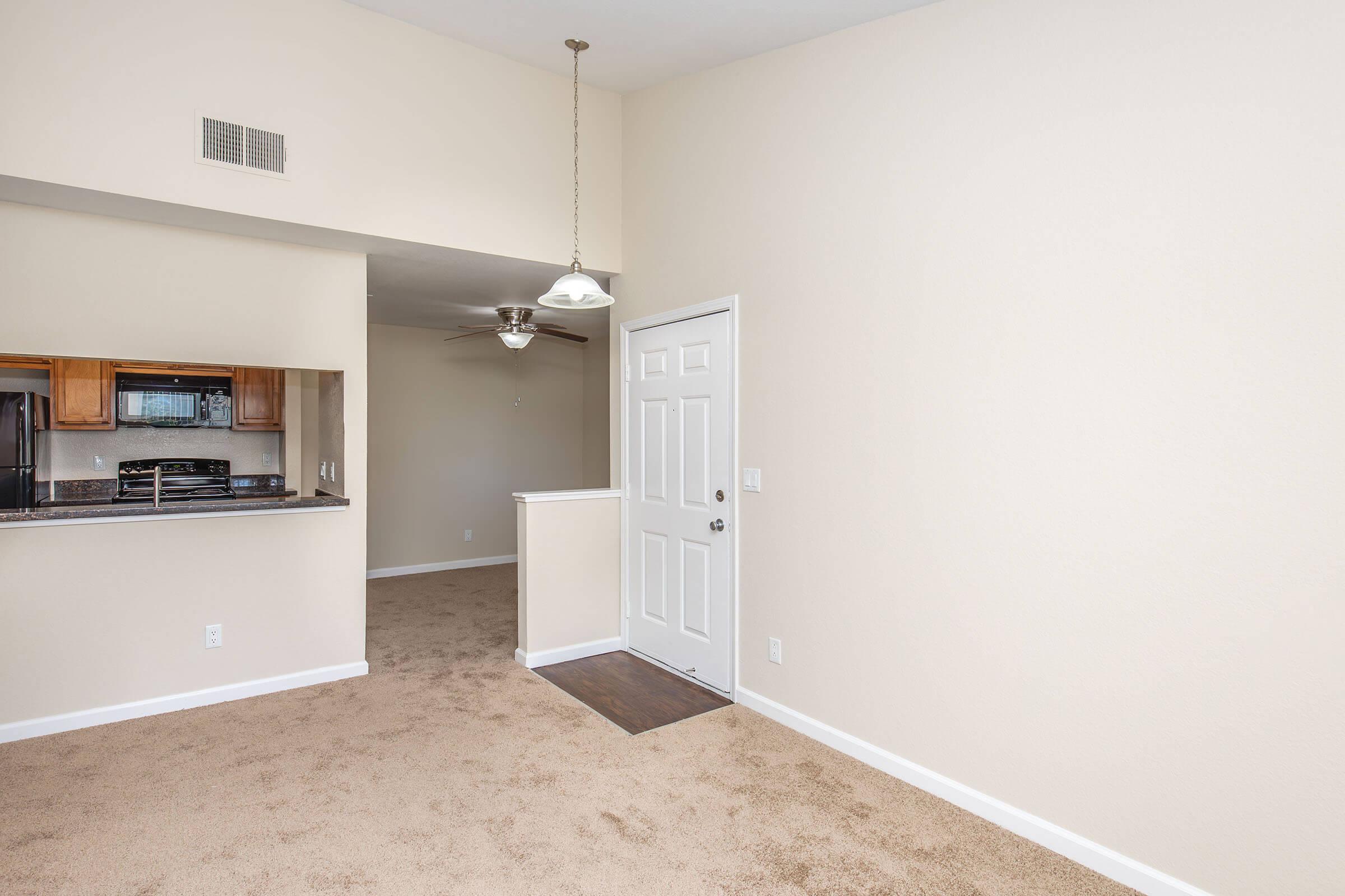 Interior view of an apartment entryway showing beige walls, carpeted floor, and a ceiling fan. A doorway leads to the kitchen area, which has wooden cabinets and an open layout. Natural light enters through the door and windows, creating a bright and inviting atmosphere. The space appears clean and unfurnished.