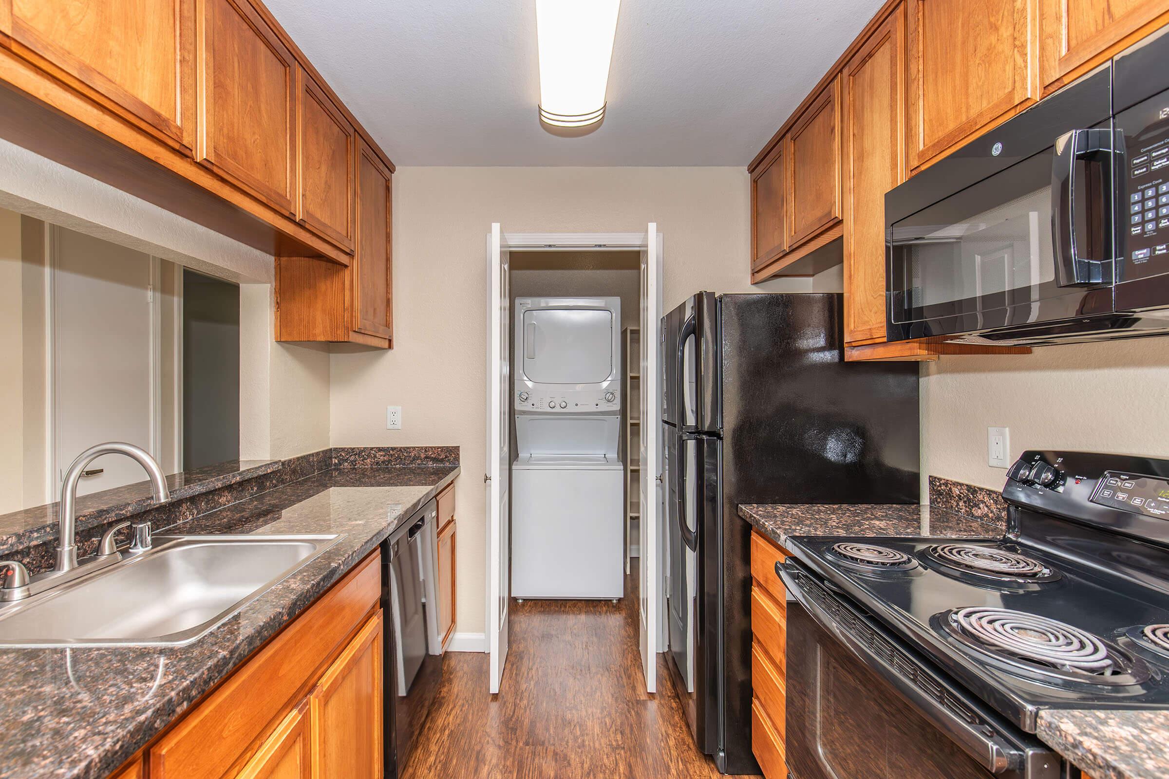 A modern kitchen featuring wooden cabinets, dark granite countertops, a stainless steel sink, and appliances including a black refrigerator, microwave, and oven. A laundry area is visible through a doorway, with a stacked washer and dryer. The floor is a warm-toned wood laminate.