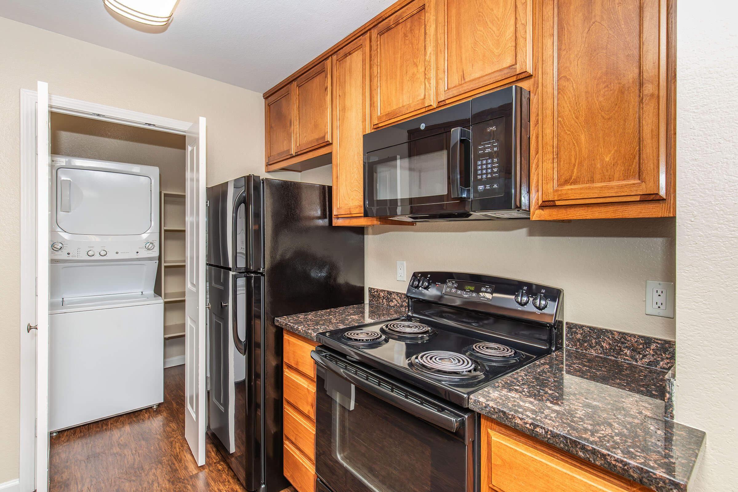 Modern kitchen featuring dark wood cabinets, black appliances including a refrigerator, oven, and microwave. A laundry area with a stacked washer and dryer is visible in the background. The countertop is dark granite, creating a sleek and functional space.