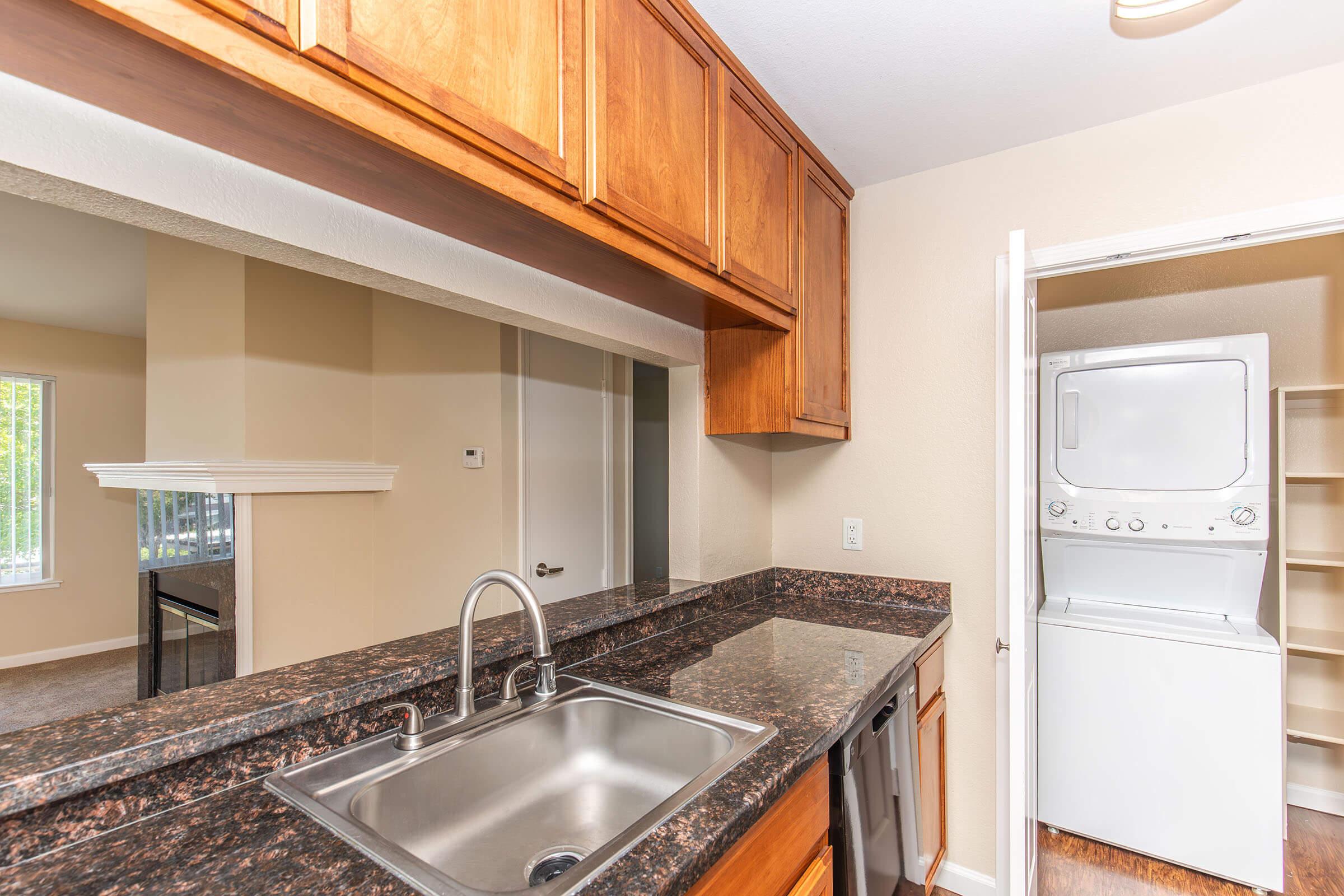 View of a modern kitchen featuring wooden cabinets above a granite countertop with a sink, an adjacent doorway leading to a laundry area with stacked washer and dryer, and a glimpse of a living space in the background with natural light.