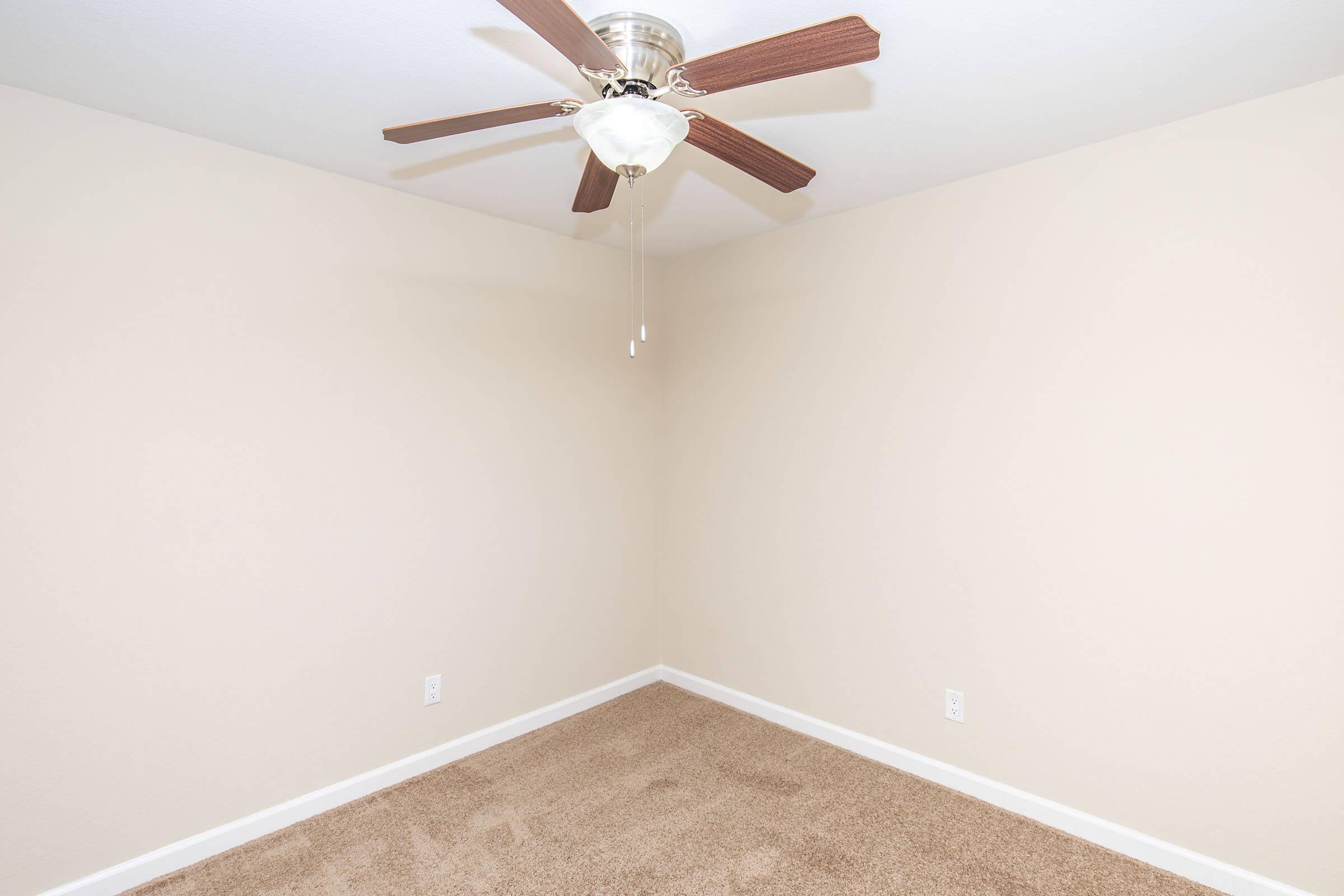 Empty room with light beige walls and a ceiling fan featuring wooden blades. The floor is covered in soft, light brown carpet. The space is unfurnished and well-lit, giving a spacious feel.