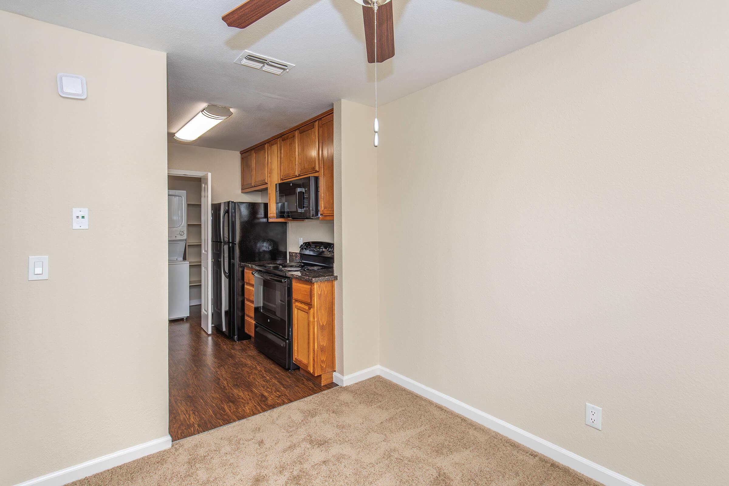 A view of a small, well-lit room with light beige walls and carpeted flooring. The room features a ceiling fan and an open doorway leading to a kitchen area with wooden cabinets, a black refrigerator, and a dark-colored stove. The overall atmosphere is cozy and modern.