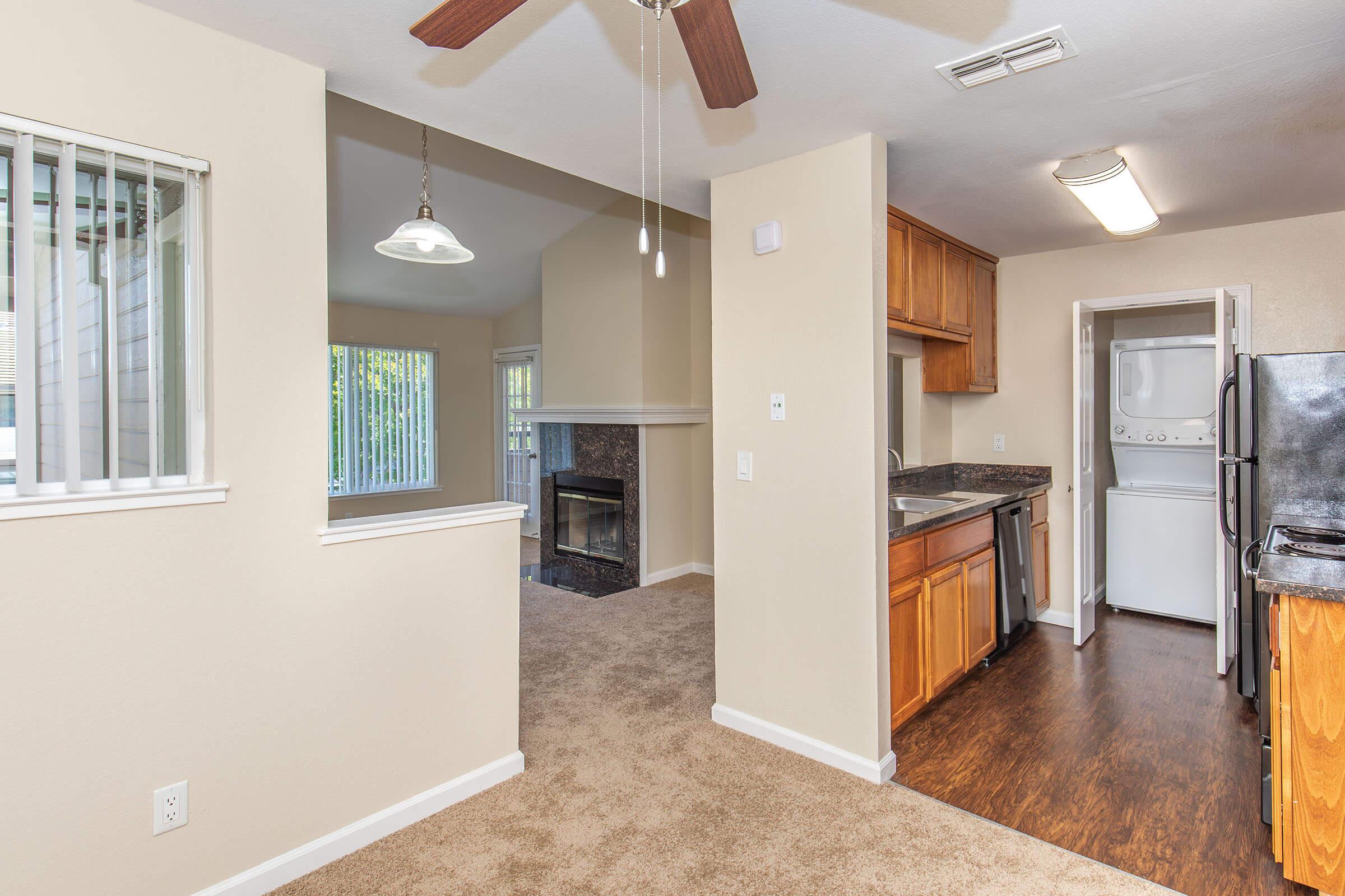 A view of an apartment interior featuring a kitchen with wooden cabinets, a countertop, and a black refrigerator. The space includes a living area with a fireplace, a ceiling fan, and a window allowing natural light. Carpeted flooring is present throughout the area.