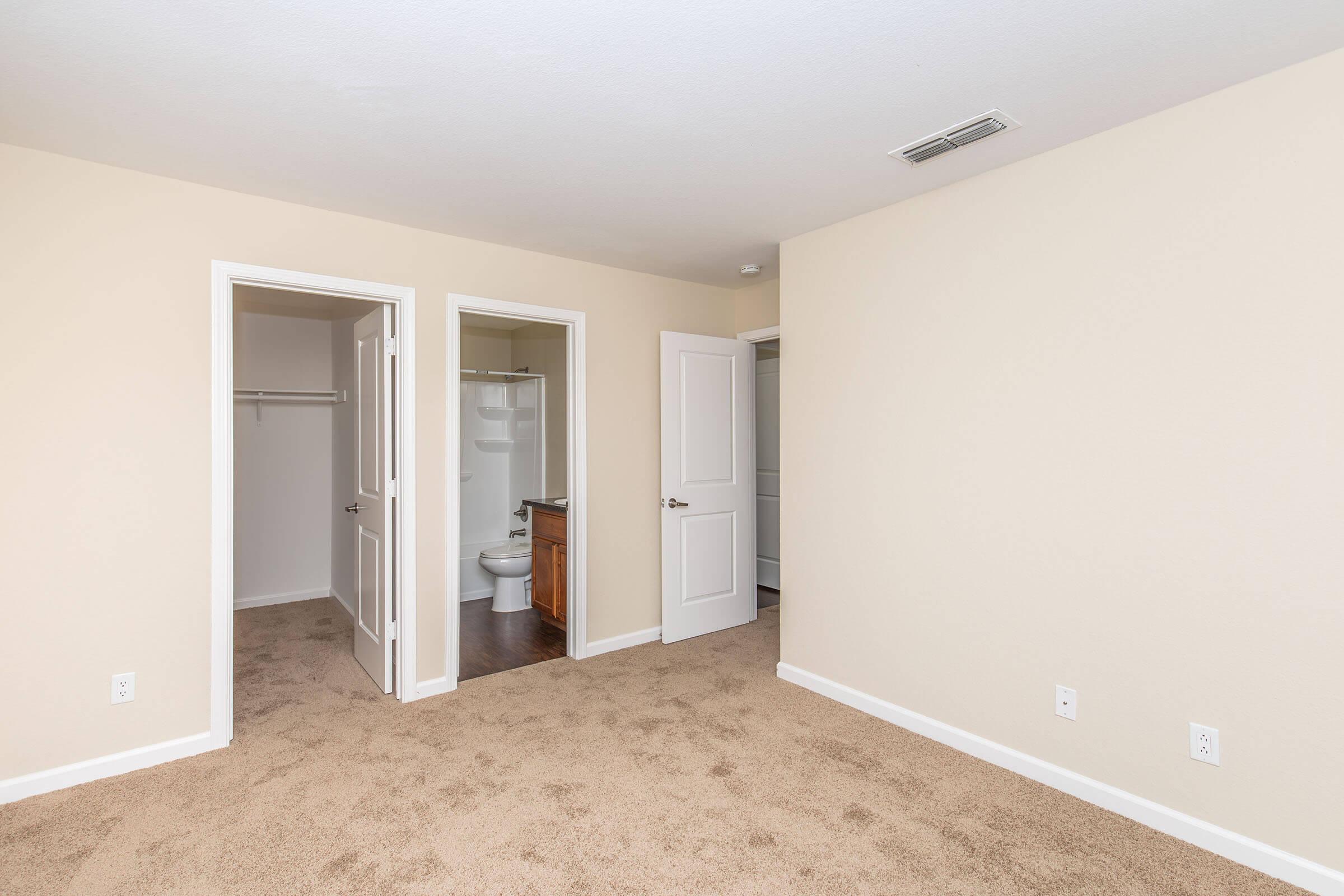 Interior of a room featuring beige walls and carpet flooring. There are three doors: one leading to a closet, another to a bathroom with a toilet, and a third leading to an adjacent room. The space appears bright and newly painted, offering a clean and simple aesthetic.