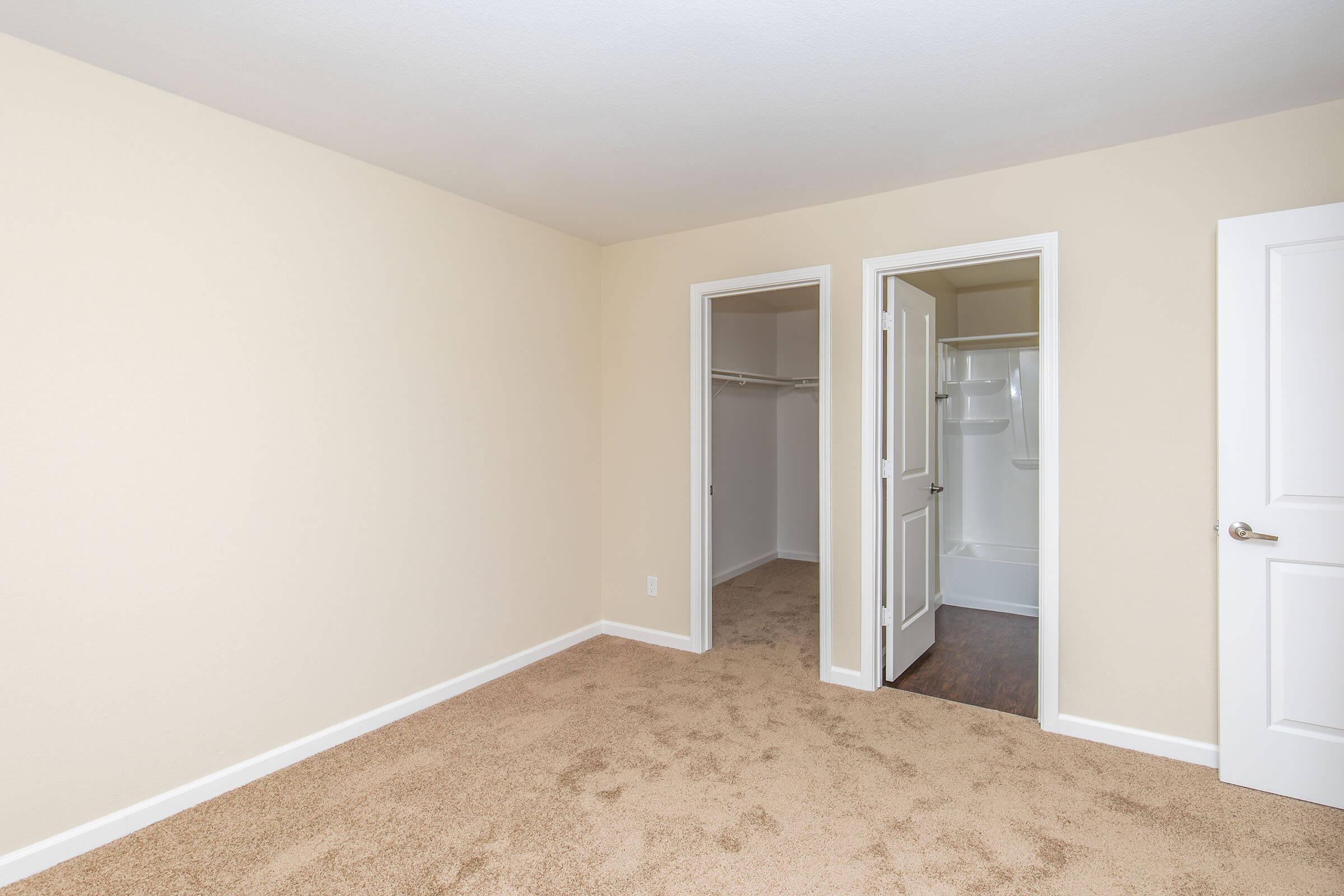 Empty room with beige walls and carpeted floor. Two white doors lead to a closet and a bathroom. The bathroom is partially visible, featuring a shower area. Overall, the space is well-lit and clean, suitable for a bedroom or guest room.