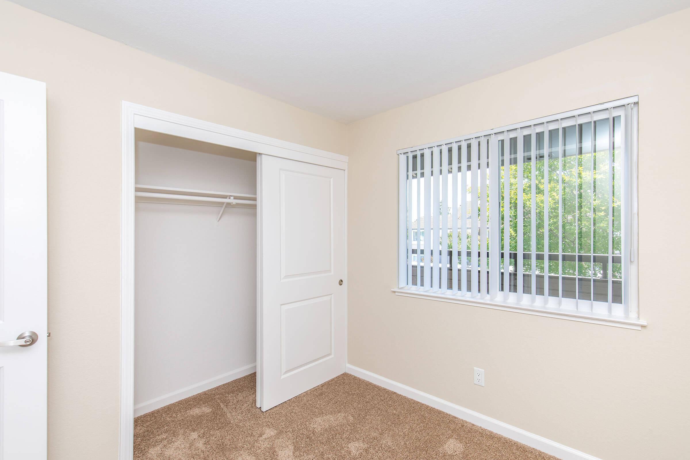 A small, well-lit bedroom featuring light beige walls and carpet. The room includes a closet with sliding doors and a window adorned with vertical blinds, allowing natural light to enter. The overall atmosphere is clean and inviting, suitable for personal use or as a guest room.