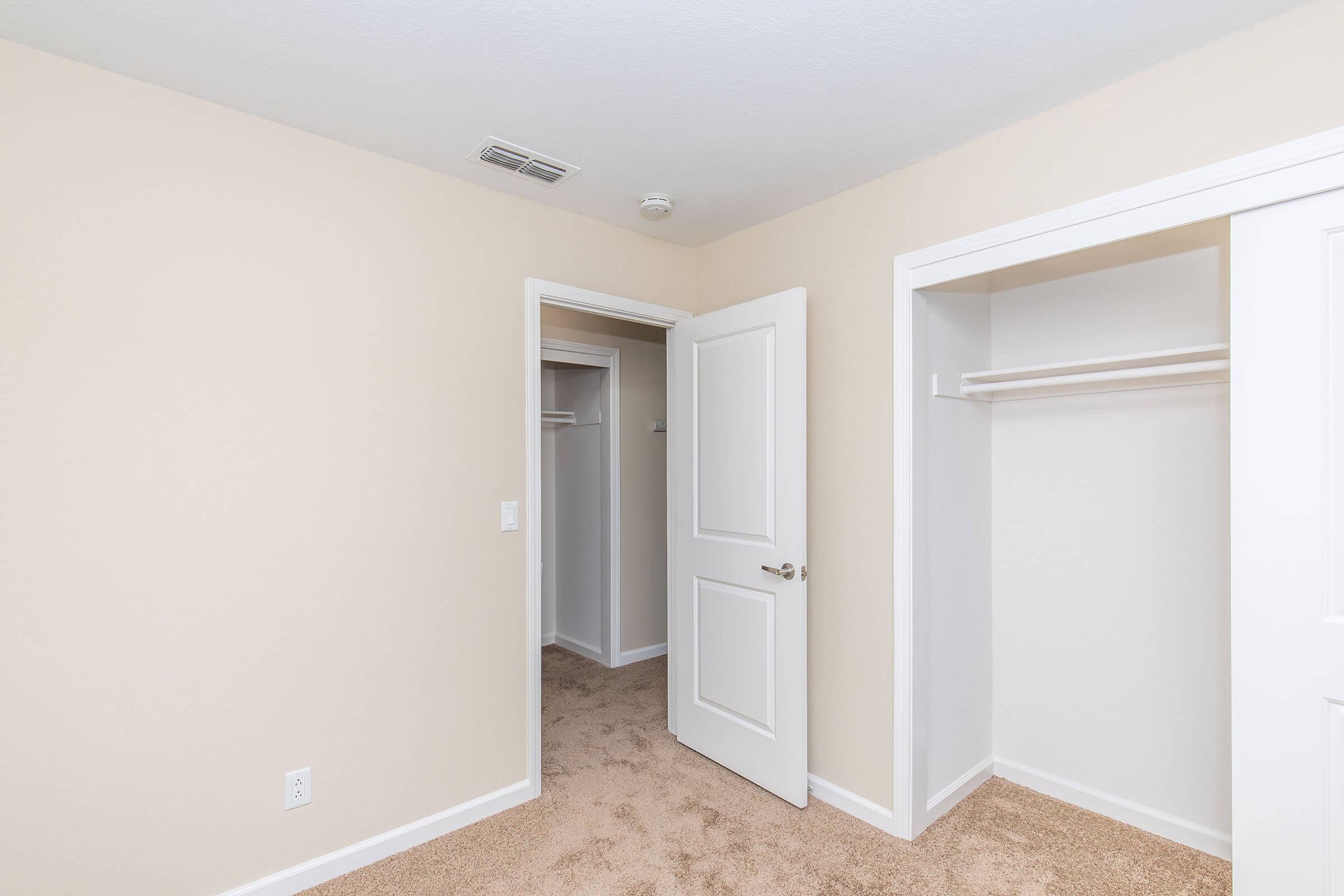 Interior view of a light-colored room featuring a beige carpet. There's an open door leading to a closet on the left, and an empty closet with a shelf on the right. The walls are painted in a soft, neutral shade, creating a bright and airy atmosphere.