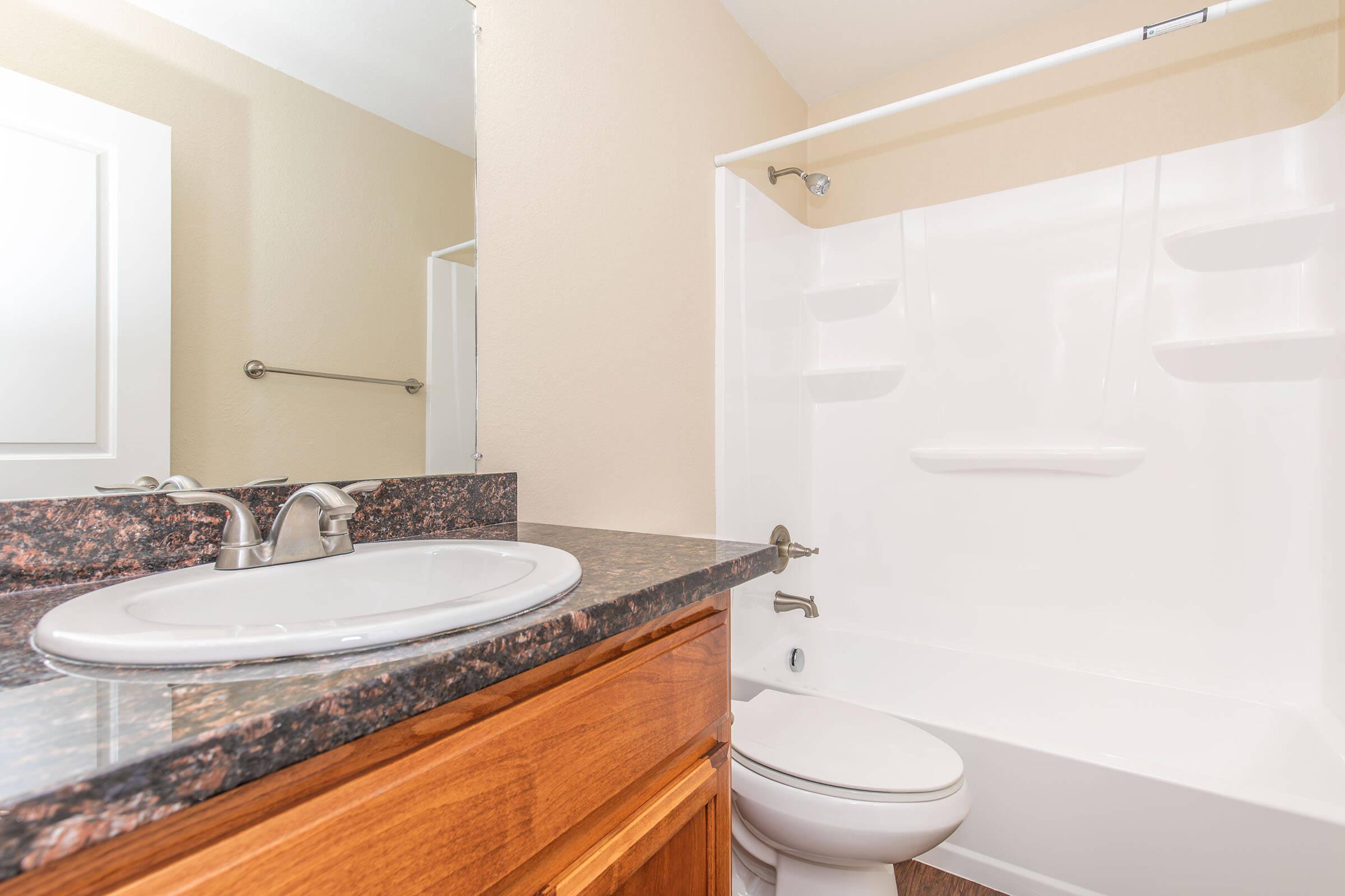 A clean bathroom featuring a white bathtub with a shower, a modern sink with a granite countertop, and a wooden cabinet. The walls are painted in a light color, and there is a mirror above the sink. A towel bar is mounted on one wall, creating an inviting and tidy space.