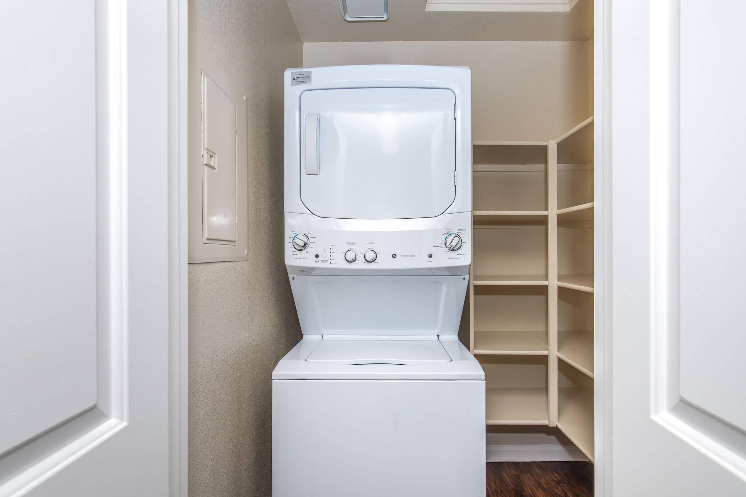 A compact laundry closet featuring a stacked washer and dryer unit in white. There are light-colored shelves on the side for storage, and the walls are painted in a neutral tone, creating a tidy and functional laundry space.
