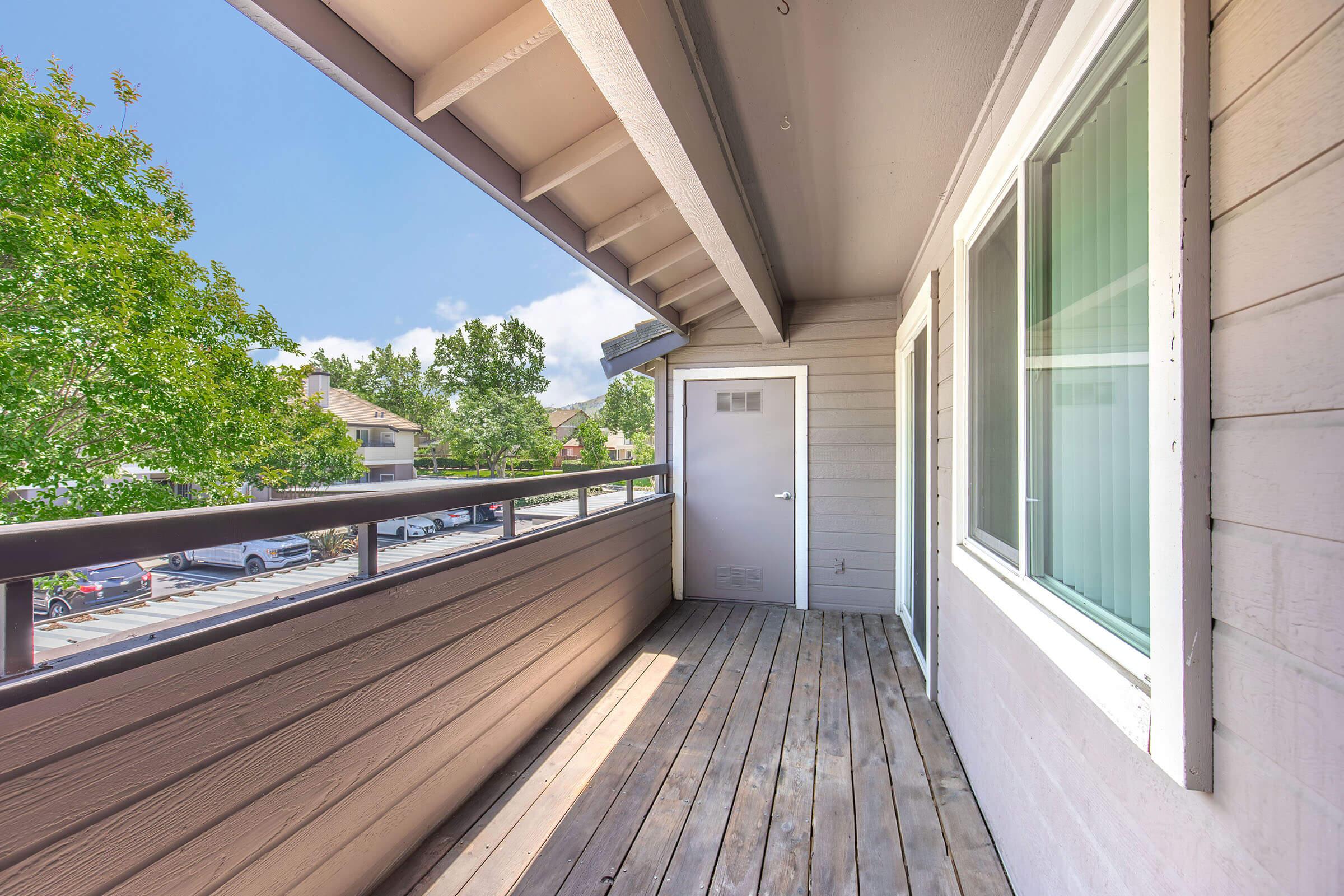 A small, wooden balcony with a railing, featuring a door on one side and large windows. The view outside includes trees and parked cars, under a clear blue sky with some clouds. The balcony has a modern design and is well-lit by natural sunlight.