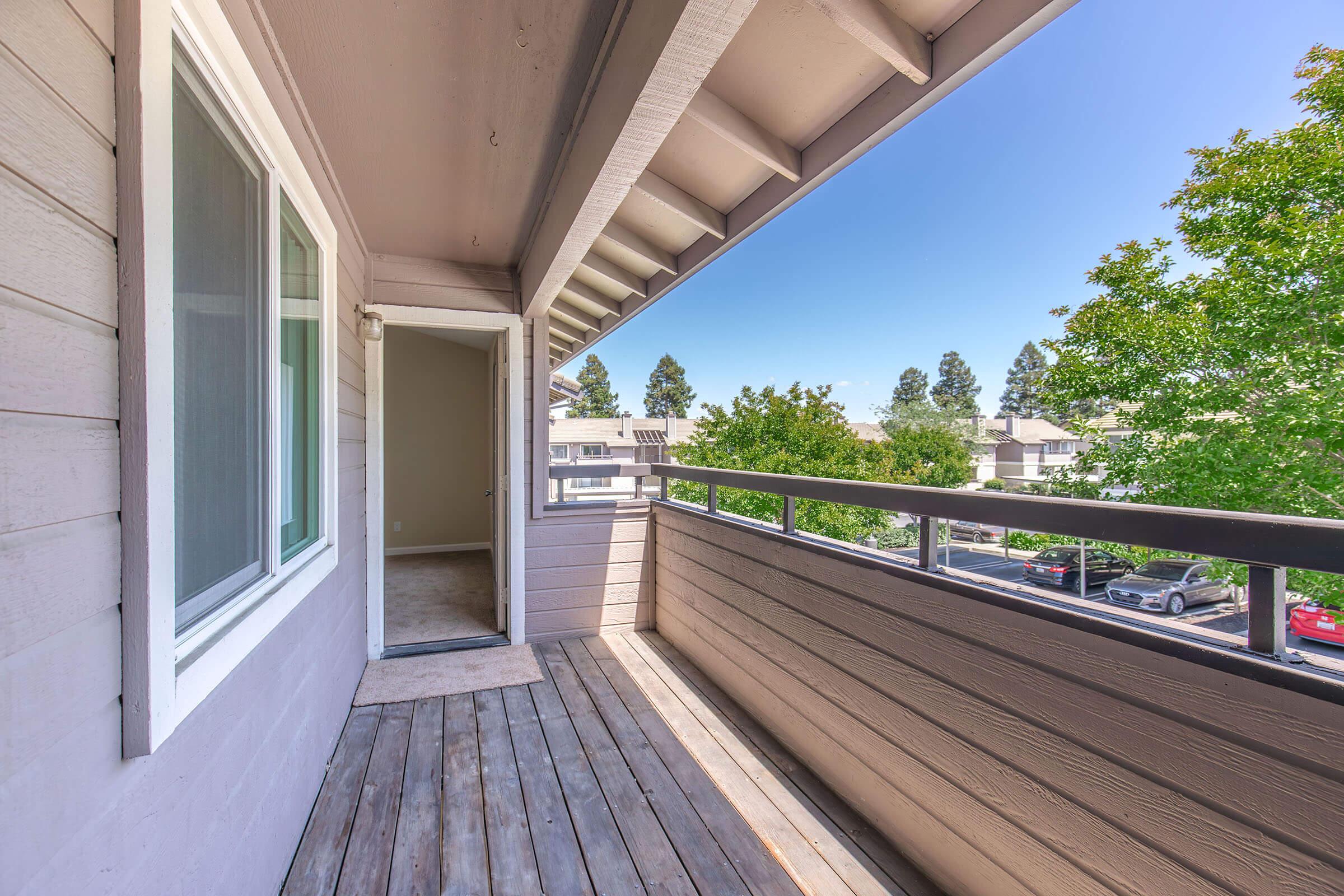 A spacious balcony with wooden flooring, featuring a railing overlooking a tree-lined area. The entrance to an interior room is visible on the left. Bright blue sky and greenery provide a serene atmosphere, ideal for outdoor relaxation.