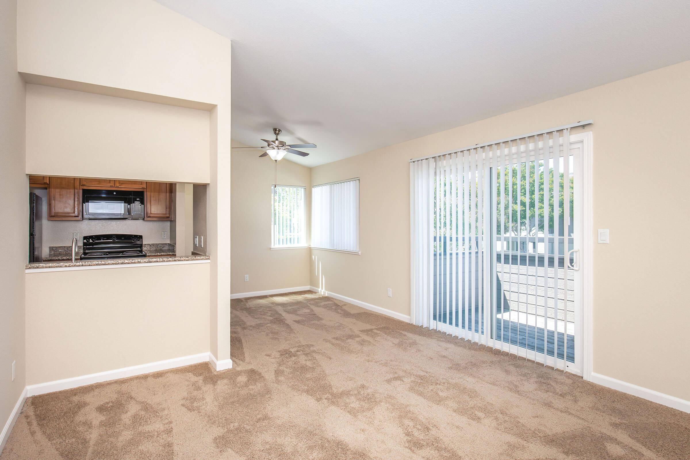 Empty room with beige walls and carpet, featuring a small kitchen area with a granite countertop and a ceiling fan. Large sliding glass doors lead to a patio outside, and there are vertical blinds on the windows allowing natural light.