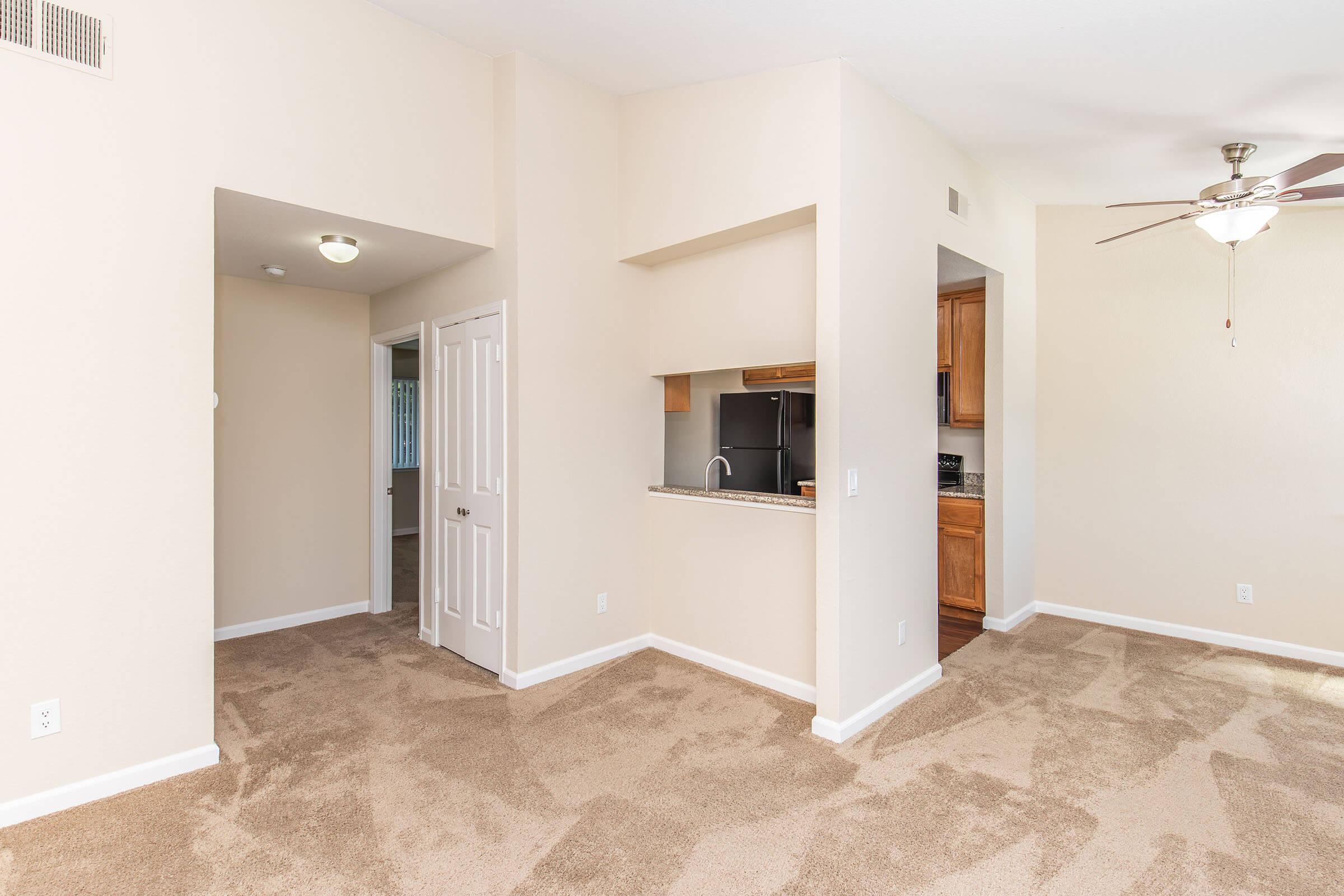 Interior view of a light-colored room featuring beige carpet flooring, a doorway leading to another room, a small kitchen area with wooden cabinets, and a ceiling fan. Natural light is visible, creating a bright and inviting atmosphere.