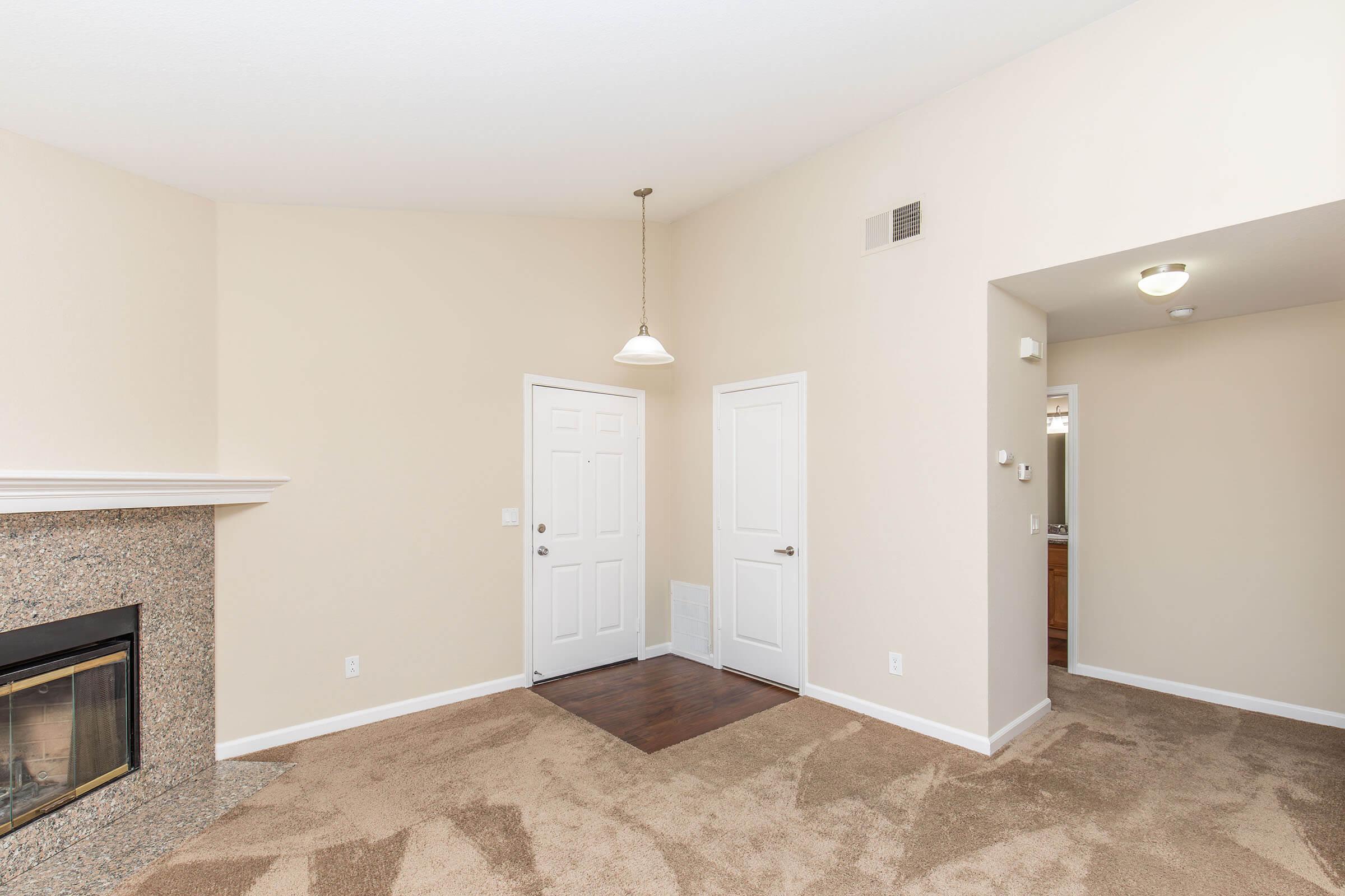 A spacious, well-lit living room featuring beige walls, plush carpet, and a decorative fireplace. There is a front door on one side and an entrance to a bathroom or another room on the opposite side. A light fixture hangs from the ceiling, adding to the inviting atmosphere.