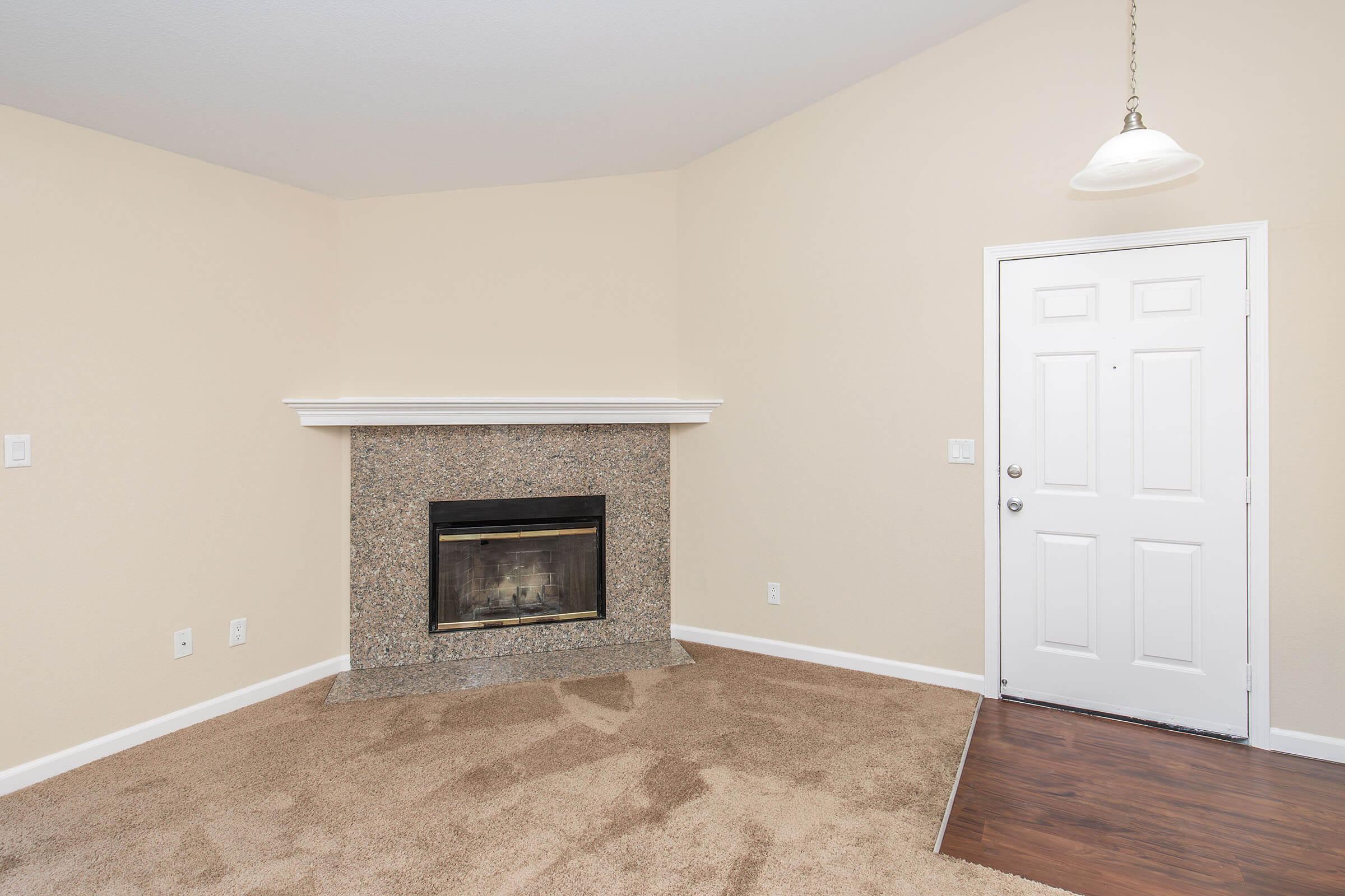 A cozy living room with beige walls and carpet, featuring a decorative granite fireplace on one side. The entrance door is white and located on the right side of the image, with a light fixture hanging from the ceiling. The space appears bright and inviting.
