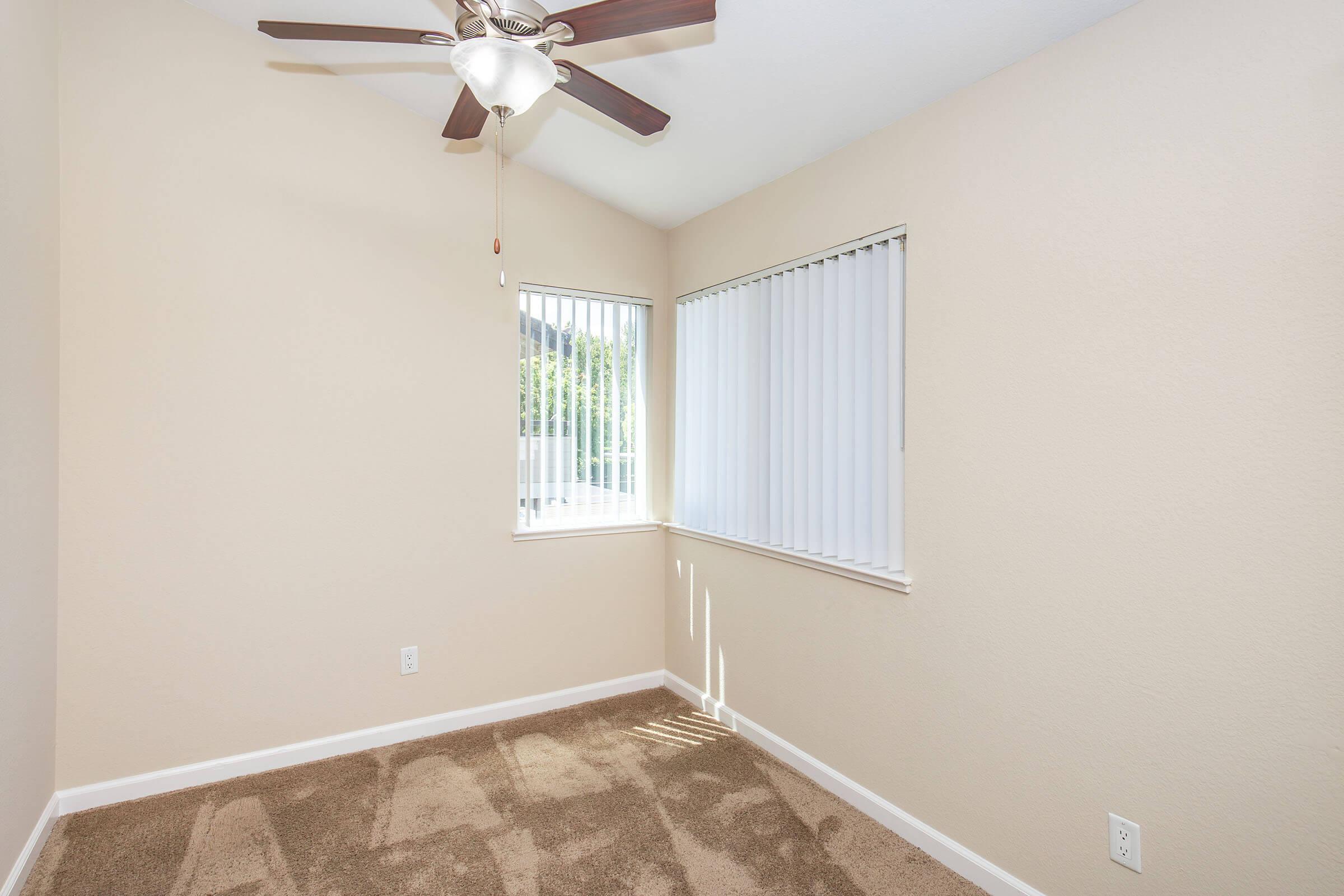 A small, empty room with light beige walls and brown carpet. There's a ceiling fan with wooden blades, and a window with vertical blinds that allows natural light to enter. The room features simple decor and has a clean, minimalist appearance.