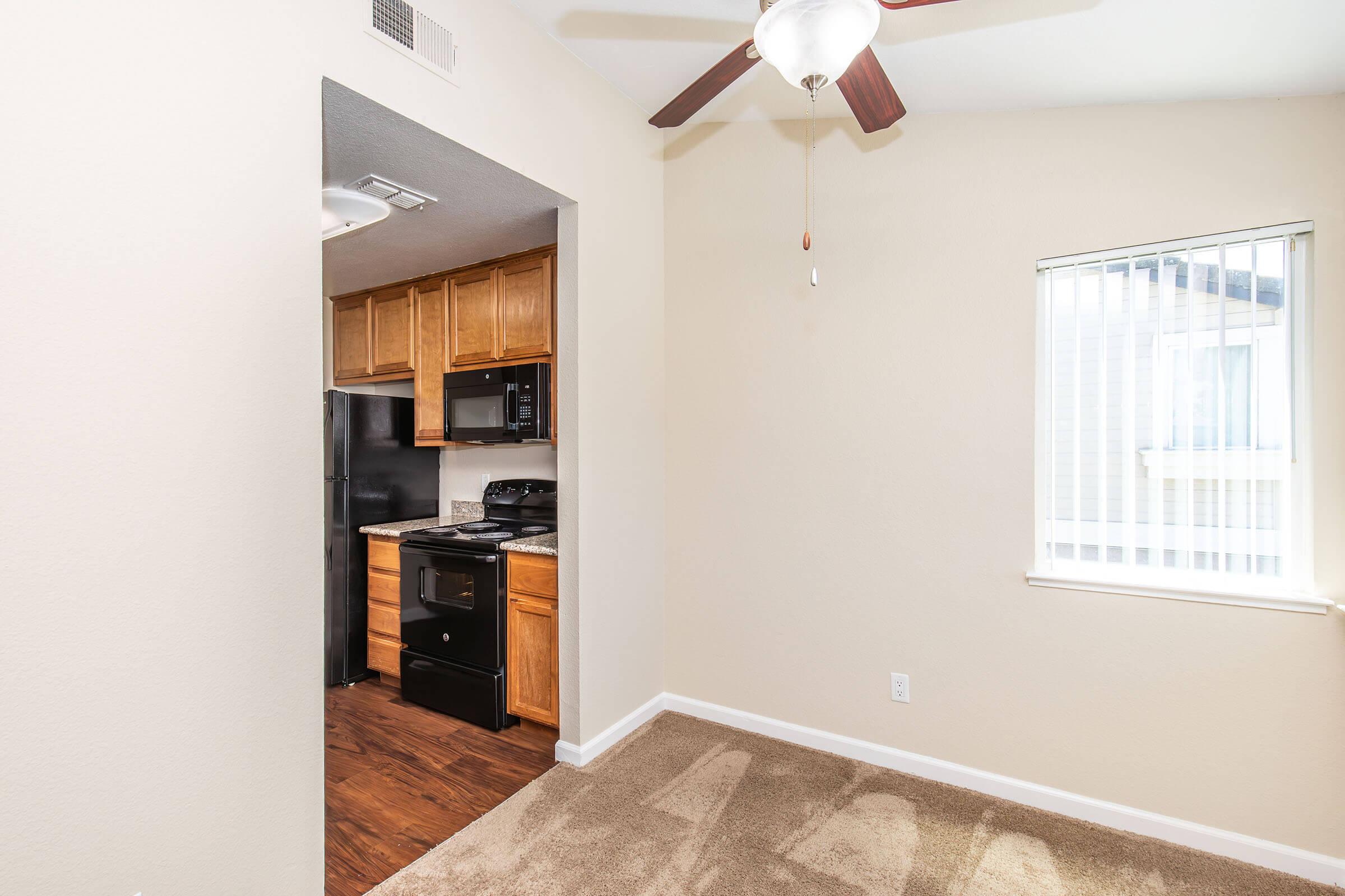 Interior view of a cozy apartment showing a small kitchen with wooden cabinets, a black refrigerator, and a stove. Nearby, a beige carpeted area and a window with blinds provide natural light, while a ceiling fan adds comfort to the space. The walls are painted a light neutral color.