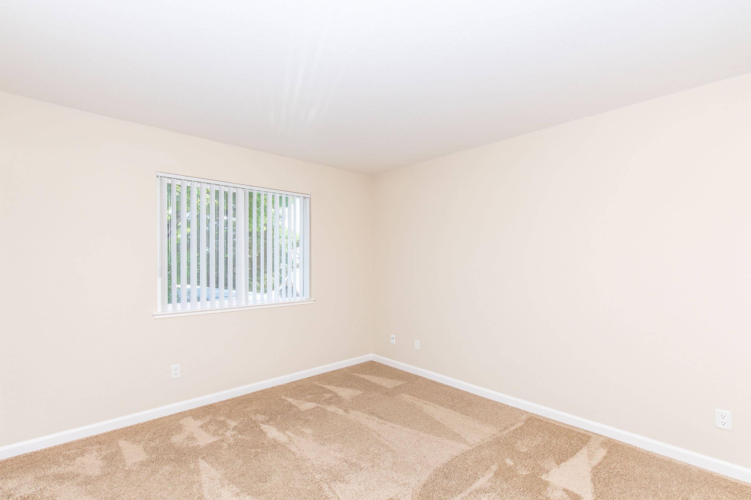 A spacious, empty room with light beige walls and carpet. A window with vertical blinds allows natural light to enter, illuminating the area. The floor is bare except for some subtle shadows from the window. The overall feel is clean and ready for furnishings.